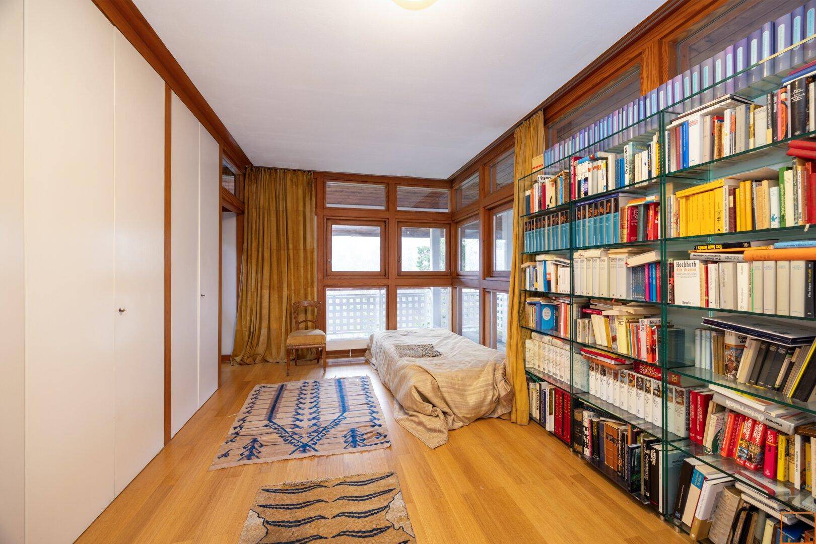 Schlafzimmer mit vielen Fenstern, Holzboden und umfangreichen Bücherregalen, die den Raum gemütlich machen.