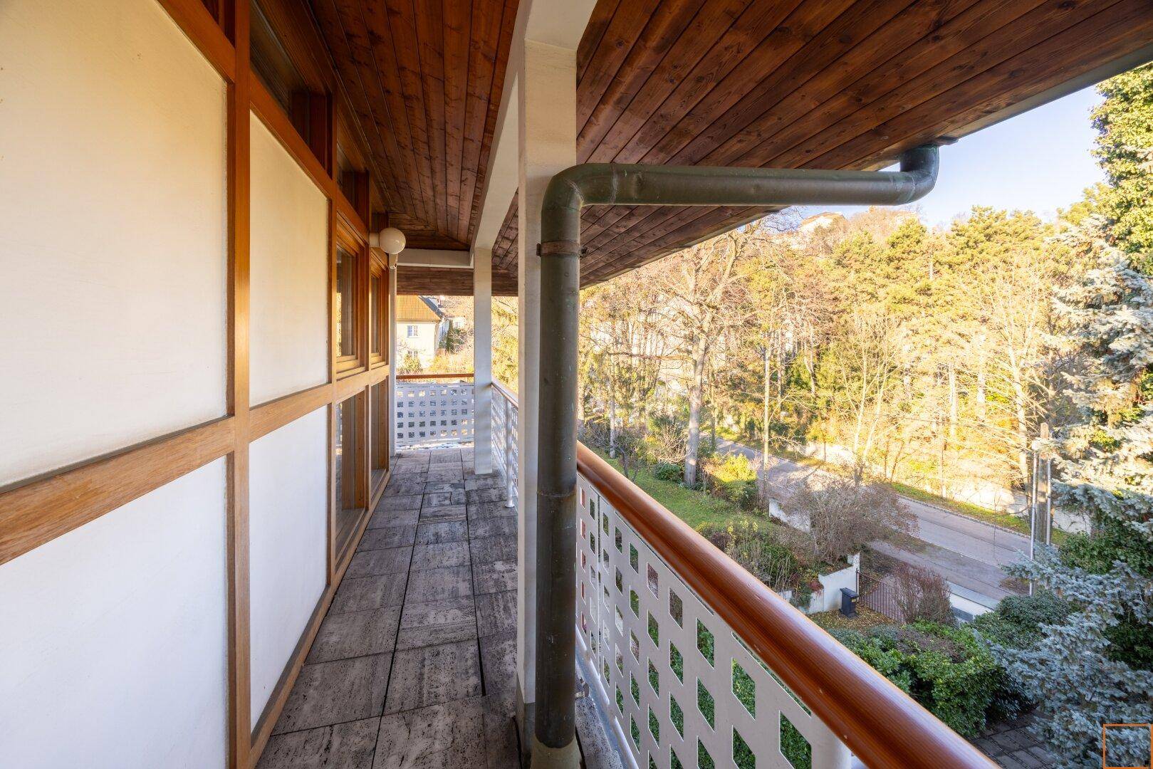 Langer Balkon mit Holzdecke und weißem Gittergeländer, der einen schönen Blick ins Grüne bietet.