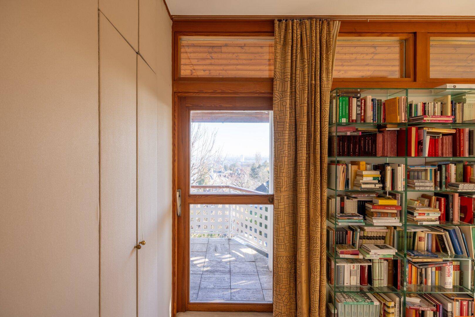 Zimmer mit Balkonzugang, Holzboden und raumhohen Bücherregalen, die viel Stauraum bieten.