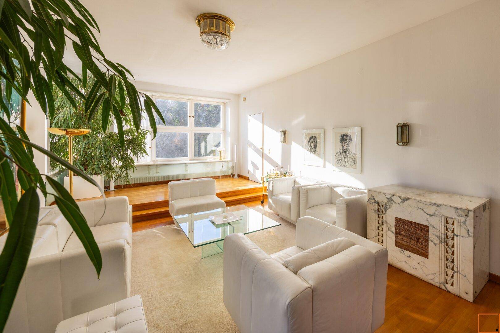 Helles Wohnzimmer mit weißen Sitzmöbeln, Holzboden und großen Pflanzen, die den Raum beleben.