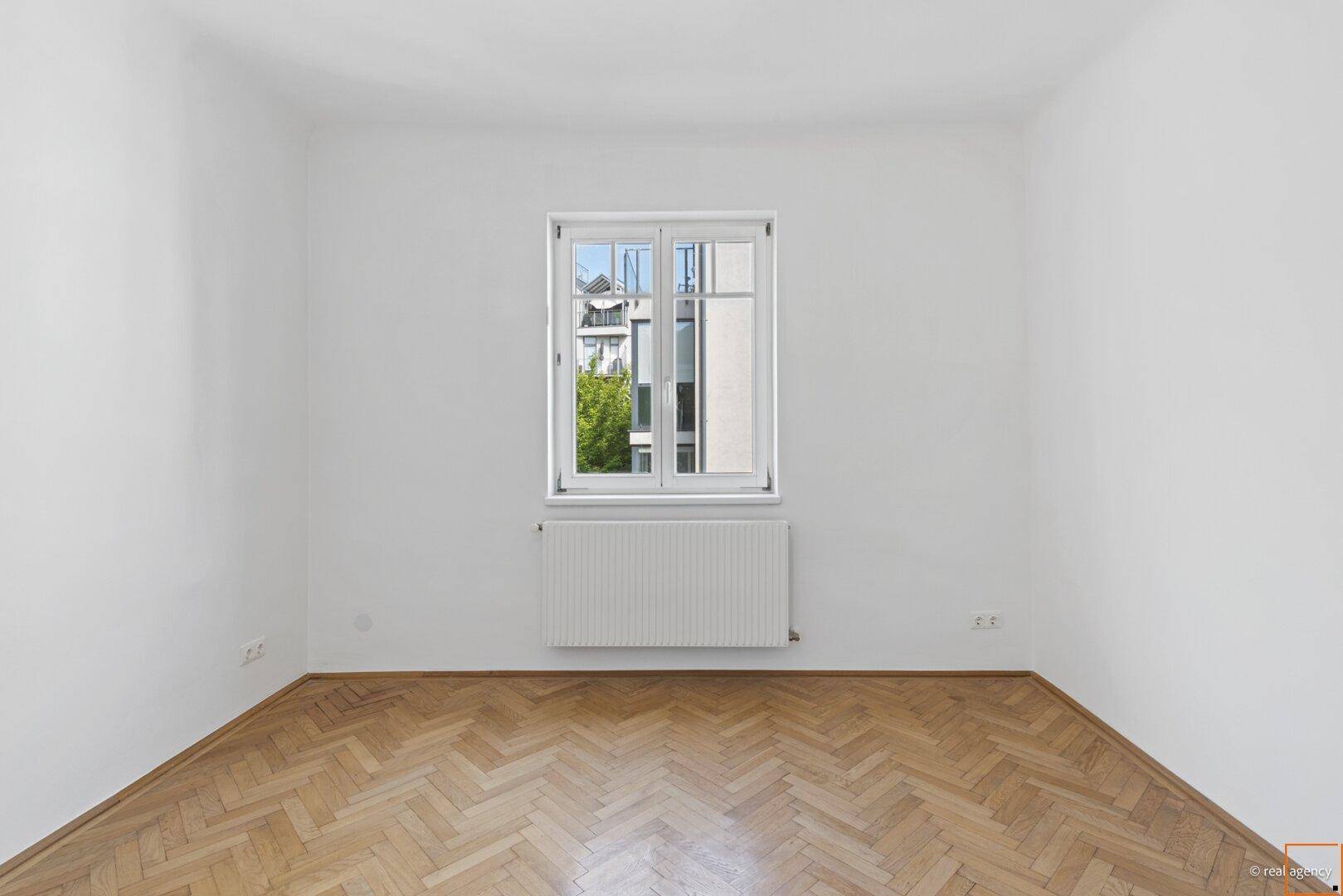 Lichtdurchflutetes Schlafzimmer mit Fischgrätparkett und einem großen Fenster mit Blick ins Grüne.