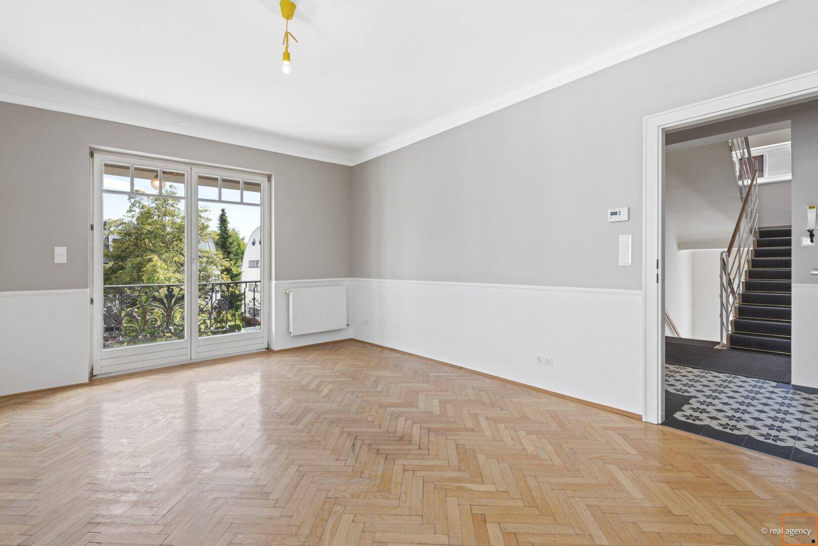 Geräumiges Wohnzimmer mit Fischgrätparkett, hellen Wänden und französischem Balkon, angrenzend an das Treppenhaus.