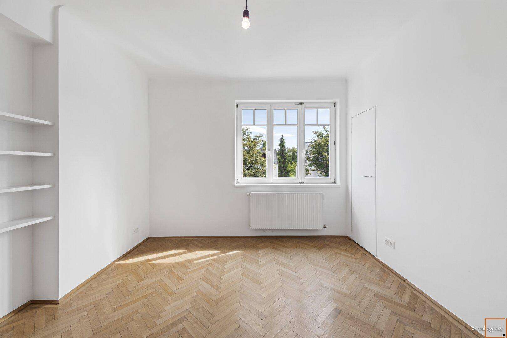Helles Schlafzimmer mit Fischgrätparkett, weißen Wänden und eingebauten Regalen neben dem Fenster.