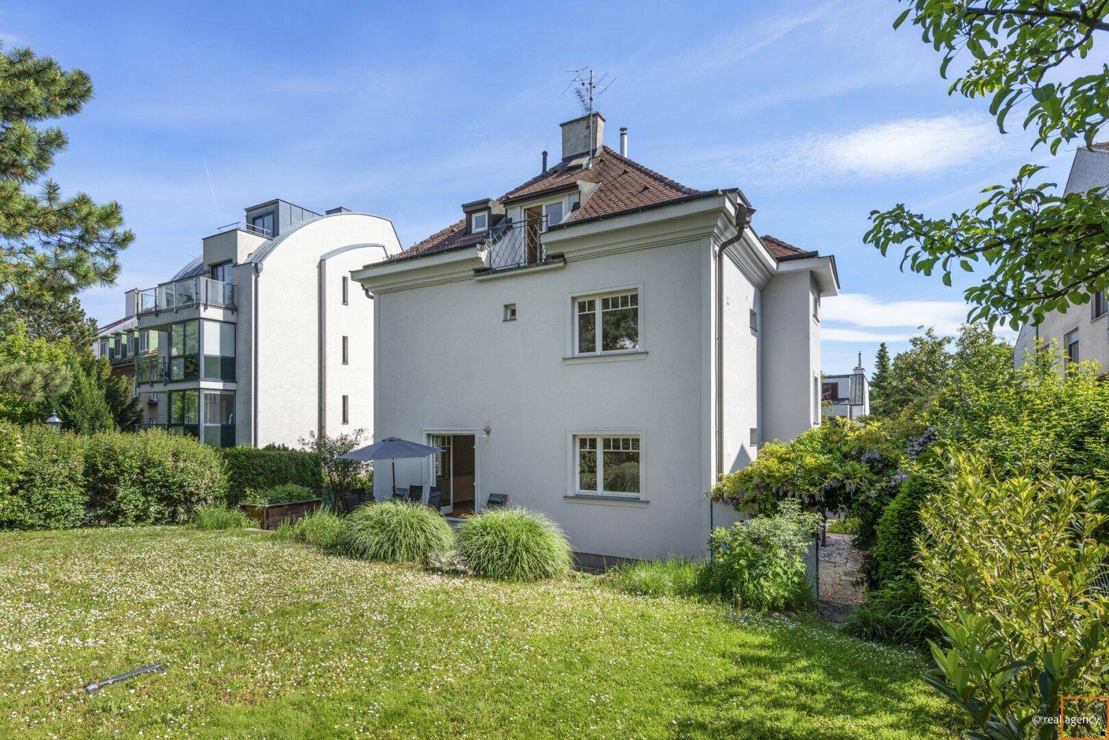 Rückansicht eines weißen Hauses mit großem, gepflegtem Garten und einer kleinen Terrasse im Freien.
