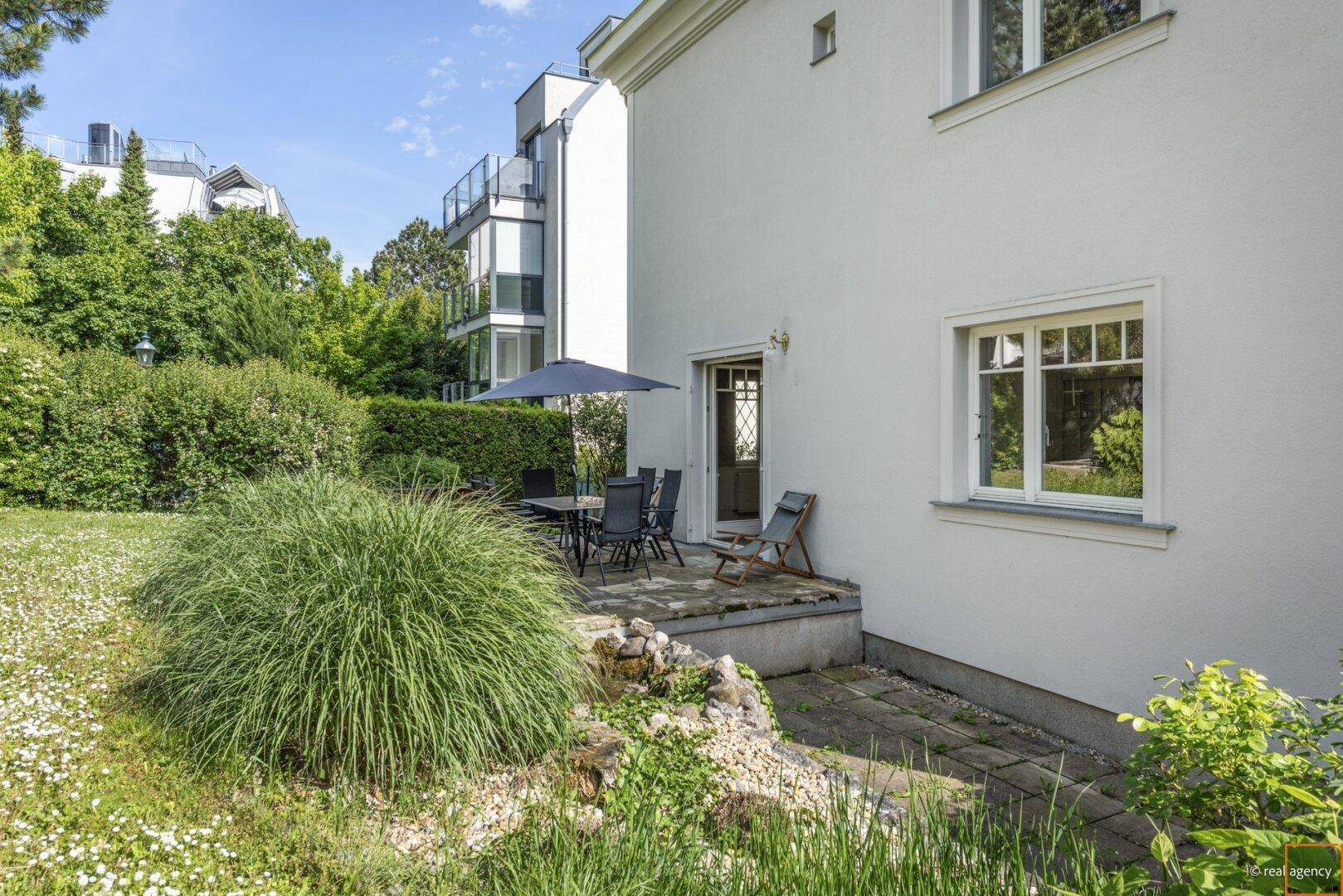Gemütliche Terrasse mit Gartenmöbeln und Sonnenschirm, umgeben von Grünflächen und Sträuchern.