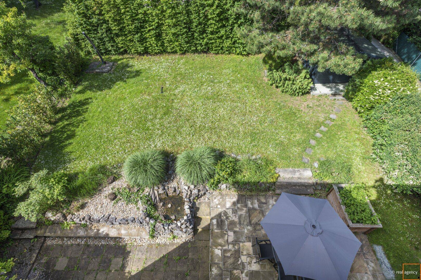 Gepflegter Garten mit Rasenfläche, Steinterrasse, Sonnenschirm und einem kleinen Teich.