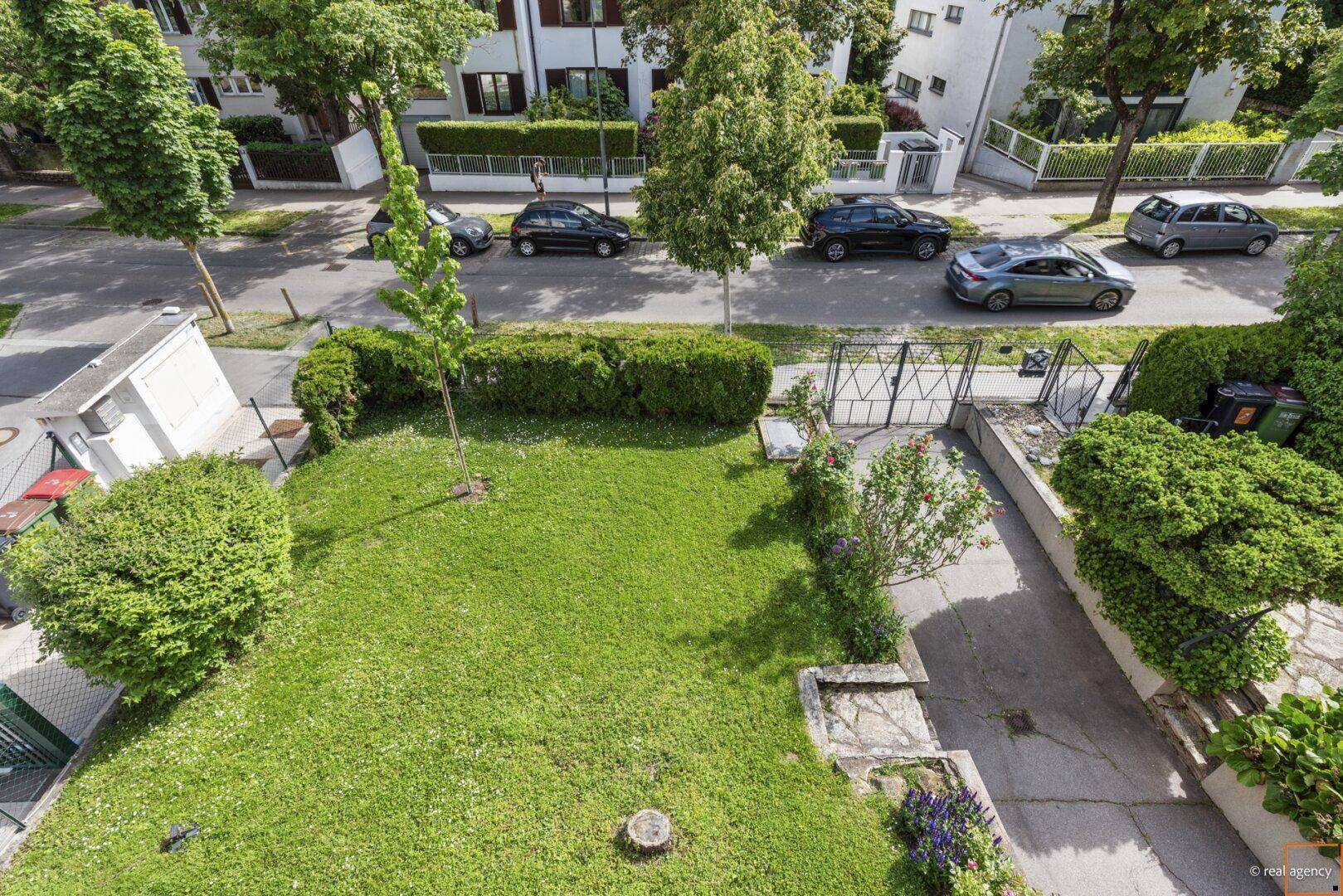 Blick von oben auf einen gepflegten Garten mit Rasenfläche und die angrenzende Straße mit parkenden Autos.
