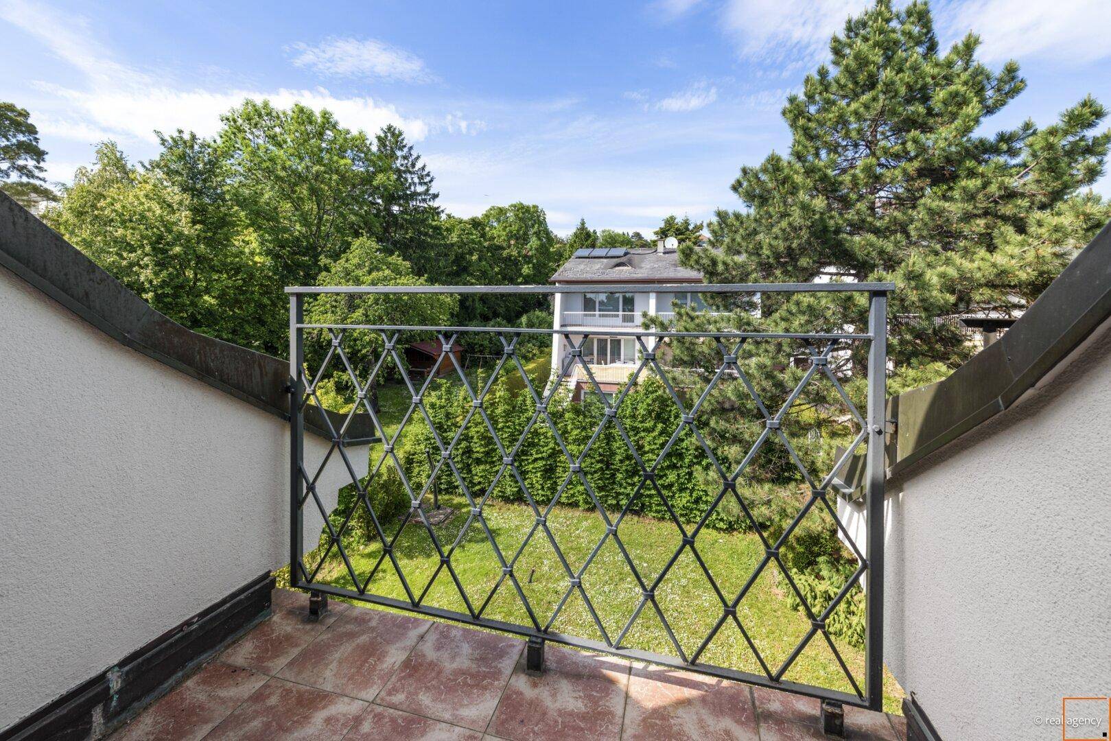 Balkon mit schmiedeeisernem Geländer und Blick ins Grüne sowie auf umliegende Häuser.