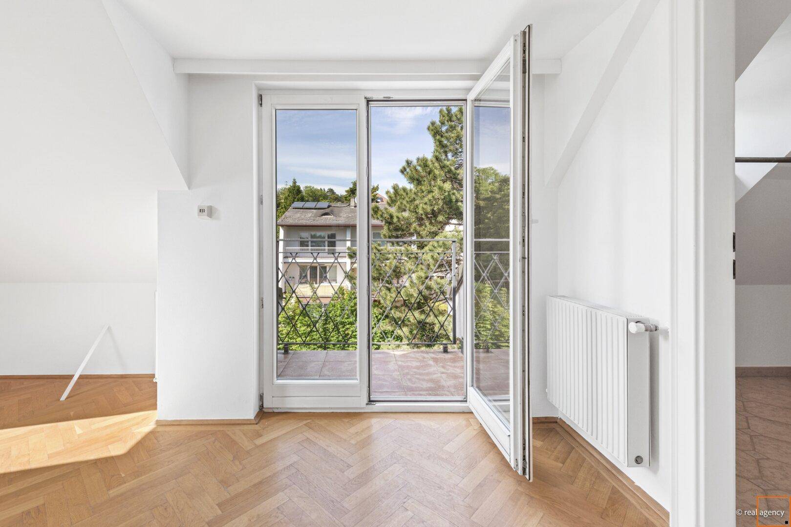 Helles Zimmer mit Fischgrätparkett, Dachschrägen und Zugang zu einem Balkon mit schmiedeeisernem Geländer.
