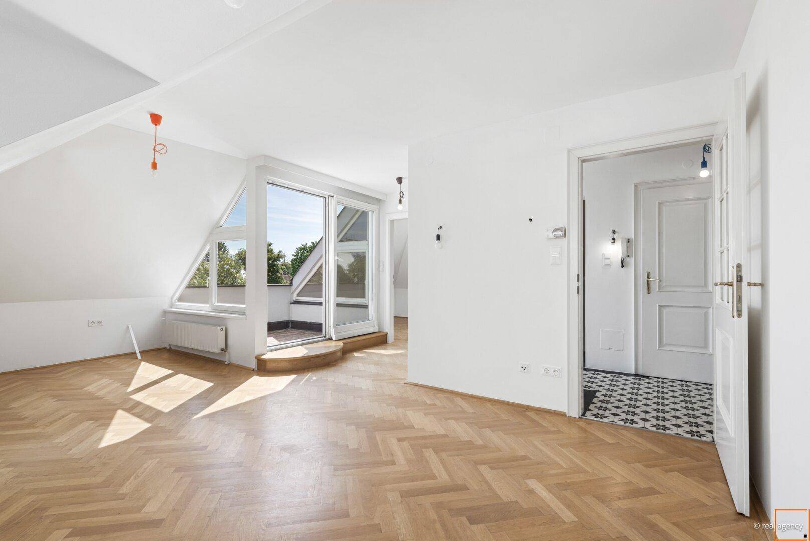 Heller Dachgeschossraum mit Fischgrätparkett, schrägen Decken und Zugang zu einem kleinen Balkon.