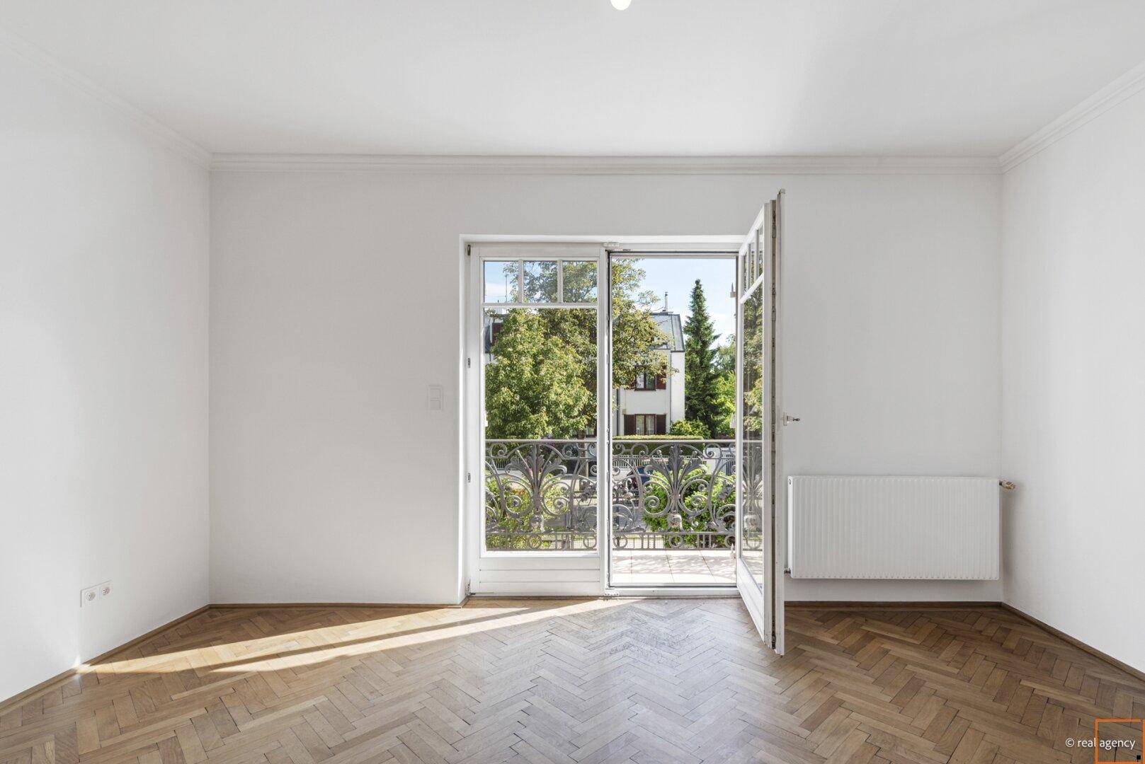 Heller Raum mit Fischgrätparkett und Zugang zu einem französischen Balkon mit schmiedeeisernem Geländer.