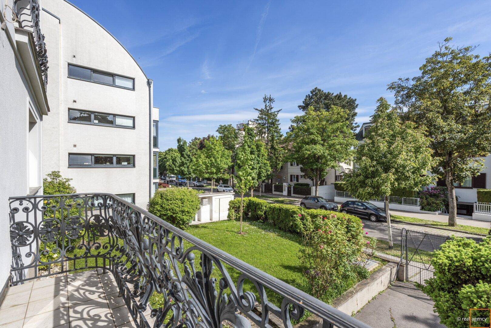 Blick vom Balkon mit schmiedeeisernem Geländer auf die grüne Straße und die umliegenden Gebäude.