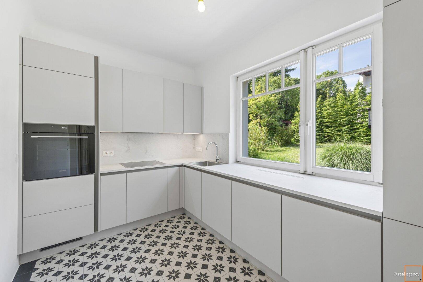 Moderne Küche mit hellgrauen Schränken, gemusterten Fliesen und großem Fenster mit Gartenblick.