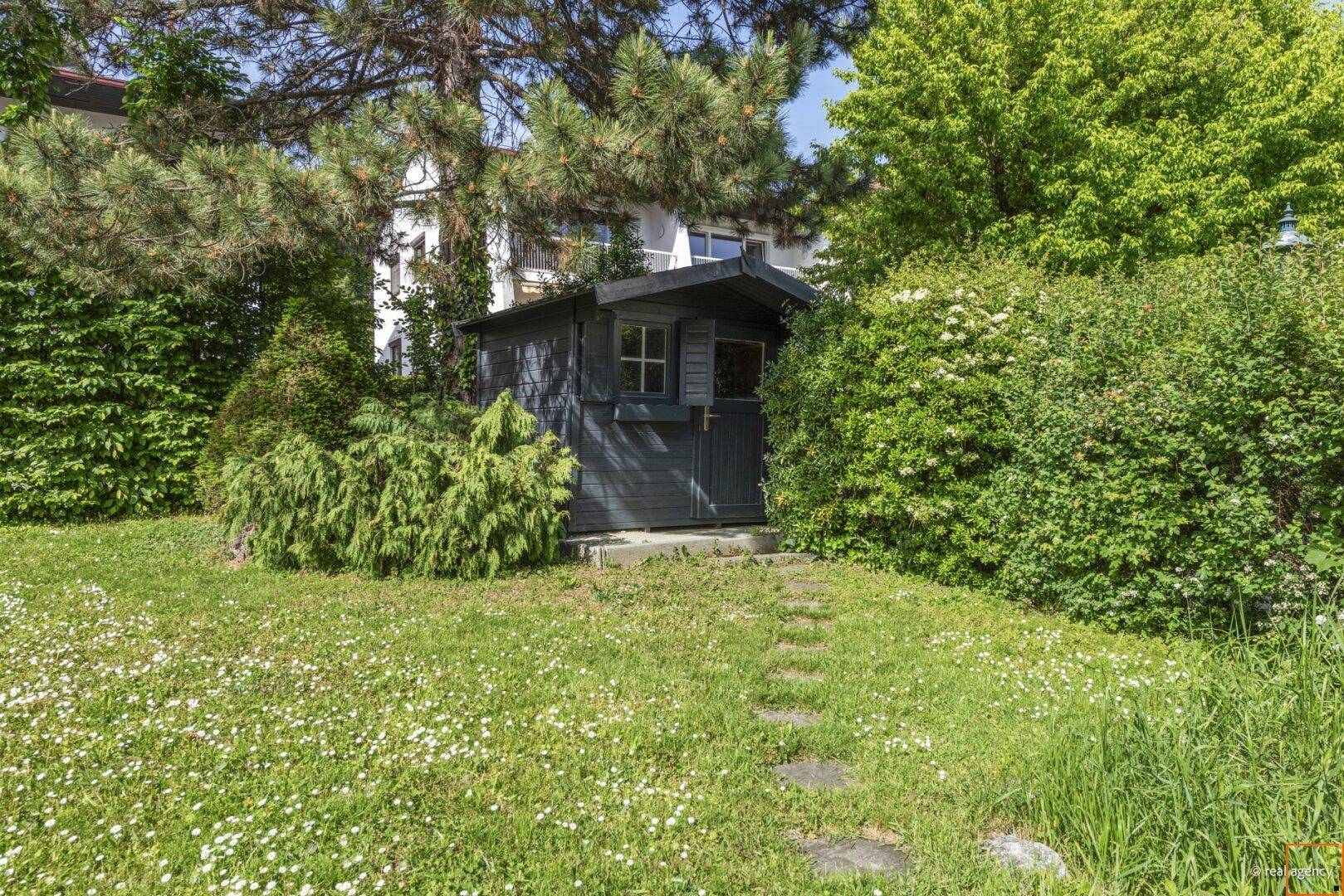 Kleiner Gartenpavillon umgeben von Bäumen und Sträuchern auf einer weitläufigen Rasenfläche.