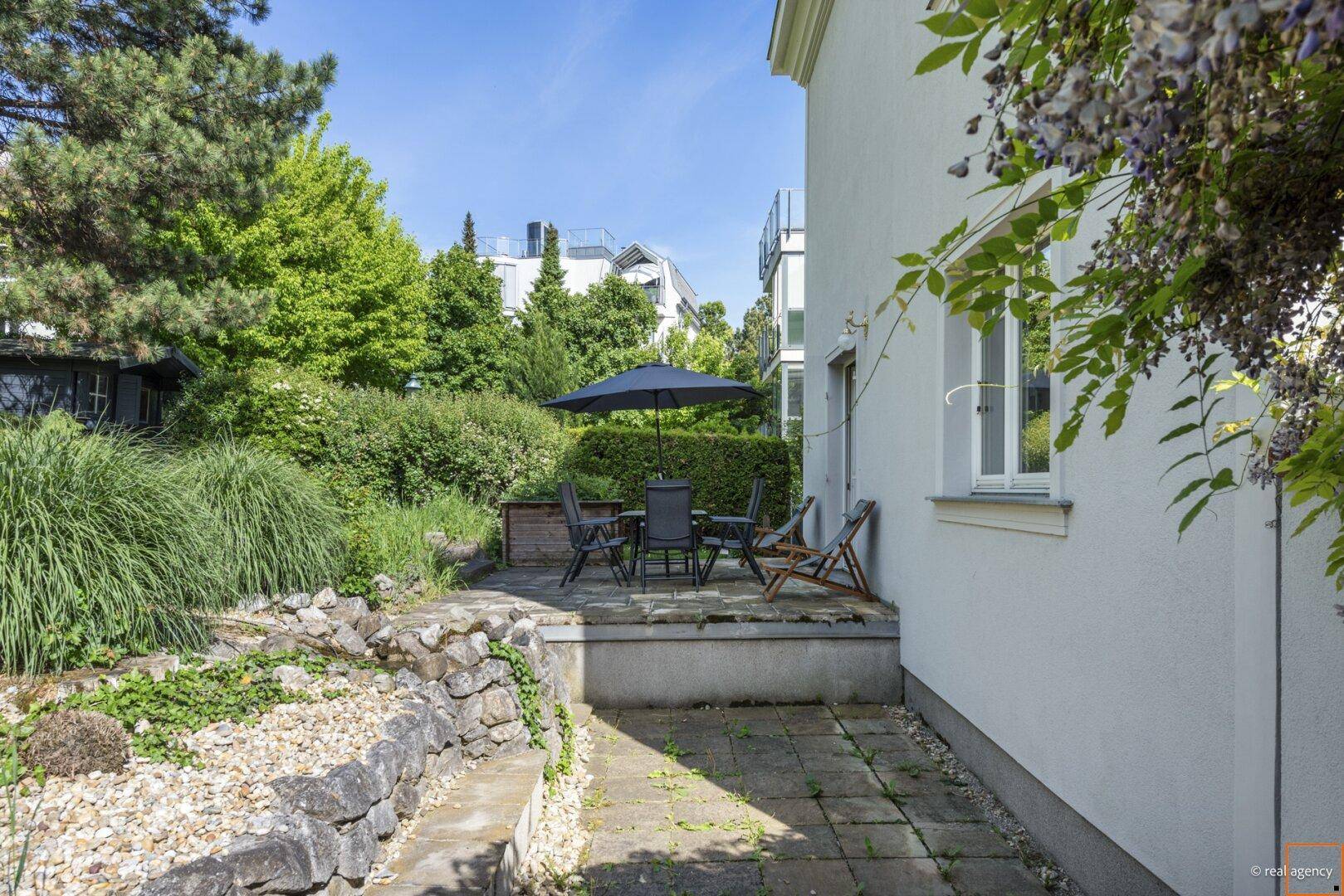 Terrasse mit Steinmauer und gepflastertem Bereich, umgeben von üppigem Grün und Sträuchern.