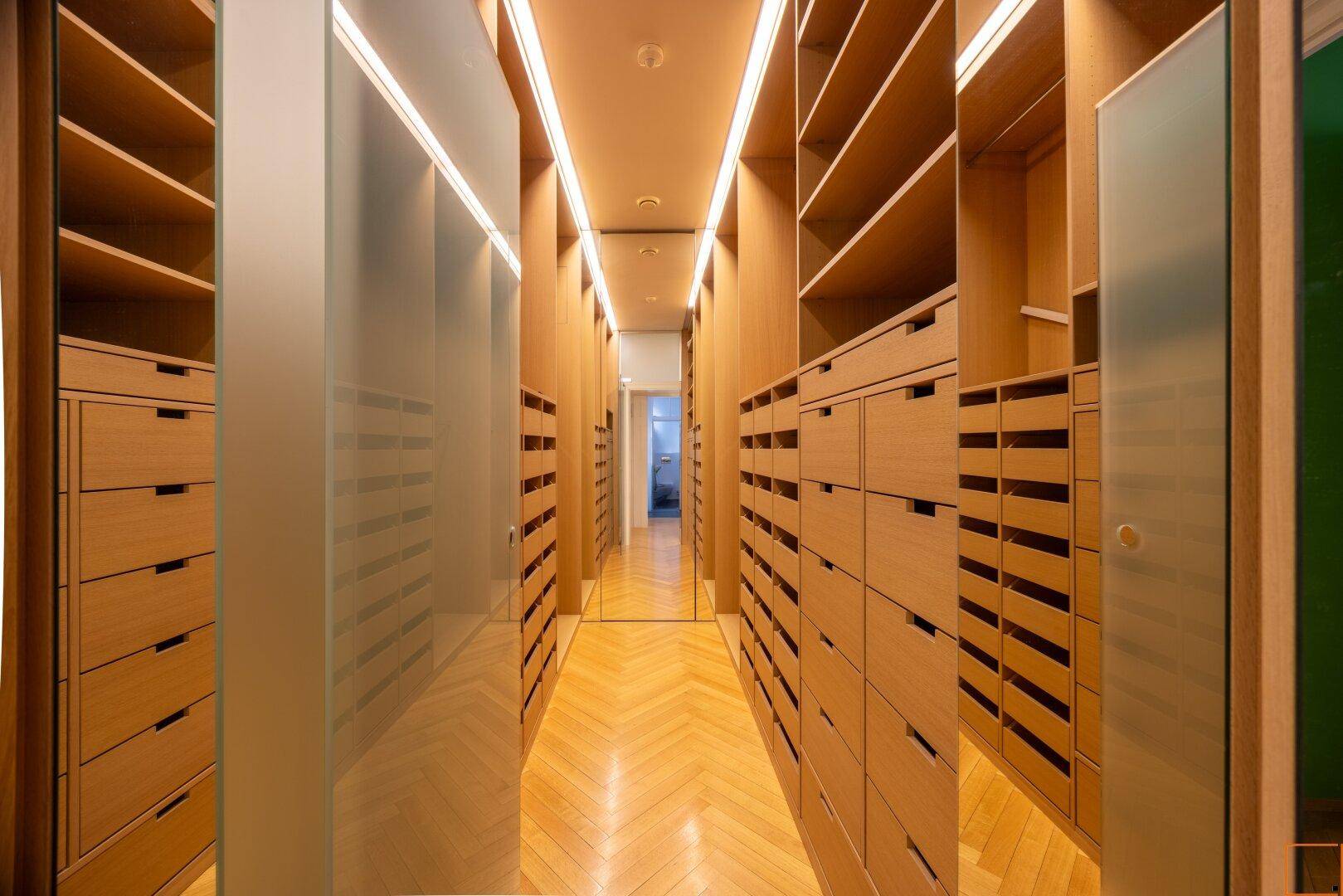 Geräumiger begehbarer Kleiderschrank mit vielen Holzregalen und Schubladen, gut beleuchtet.
