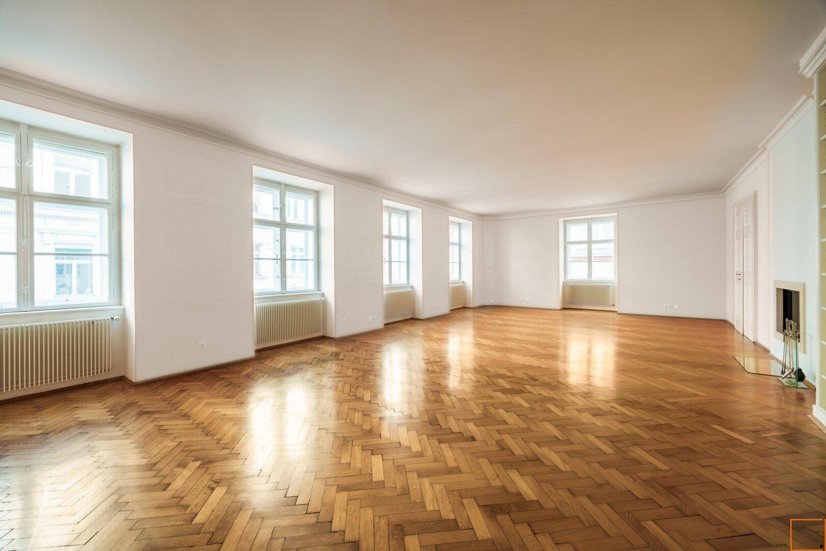 Geräumiges Wohnzimmer mit Fischgrätparkett, weißen Wänden und vielen Fenstern.