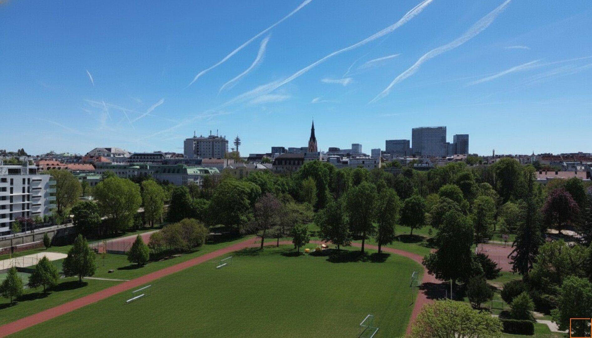Luftaufnahme einer Stadtlandschaft mit ausgedehnten Grünflächen und Sportanlagen unter blauem Himmel.