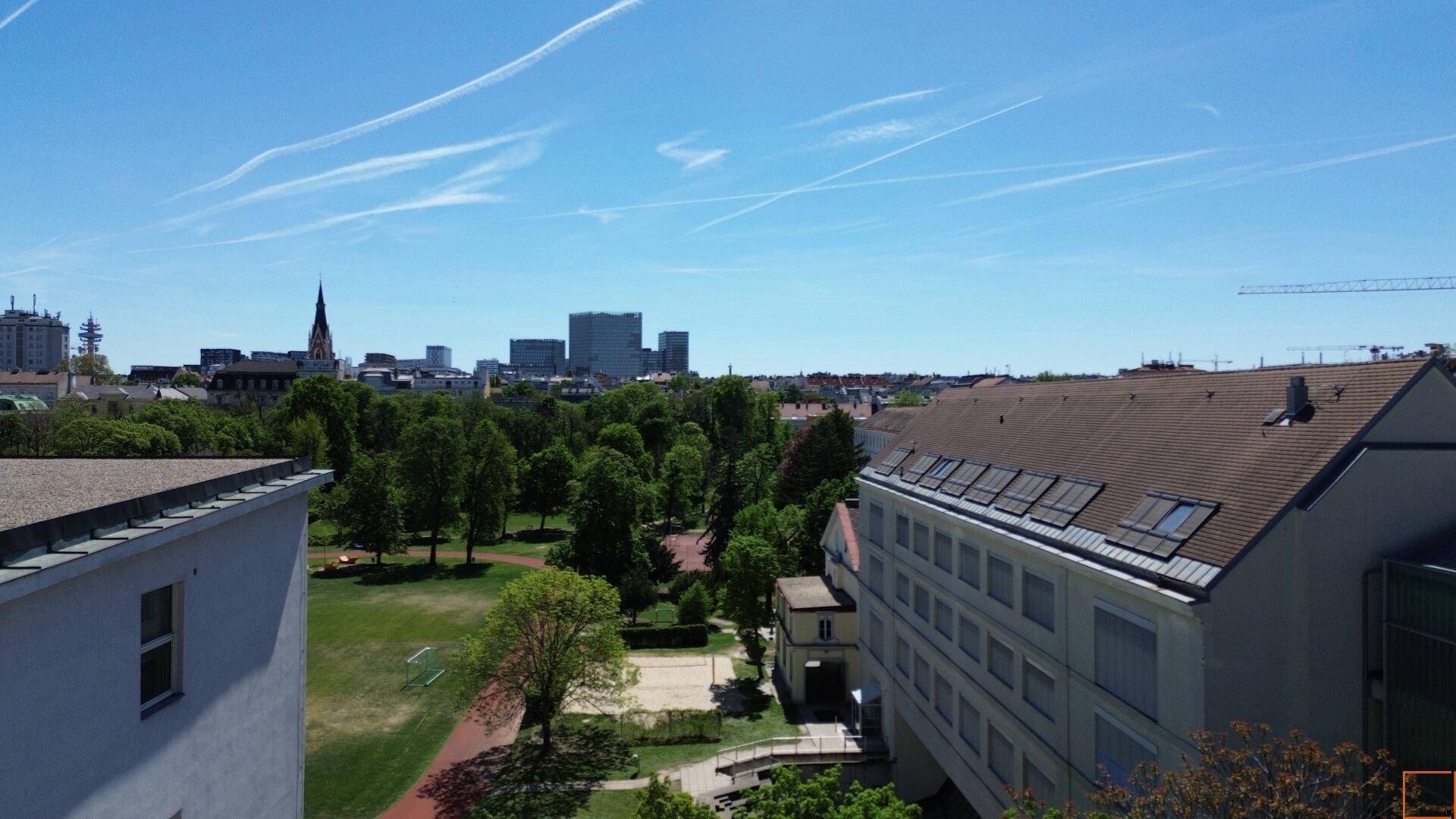 Panoramablick über eine Stadt mit grünen Parkanlagen, Wohngebäuden und einer markanten Skyline unter blauem Himmel.