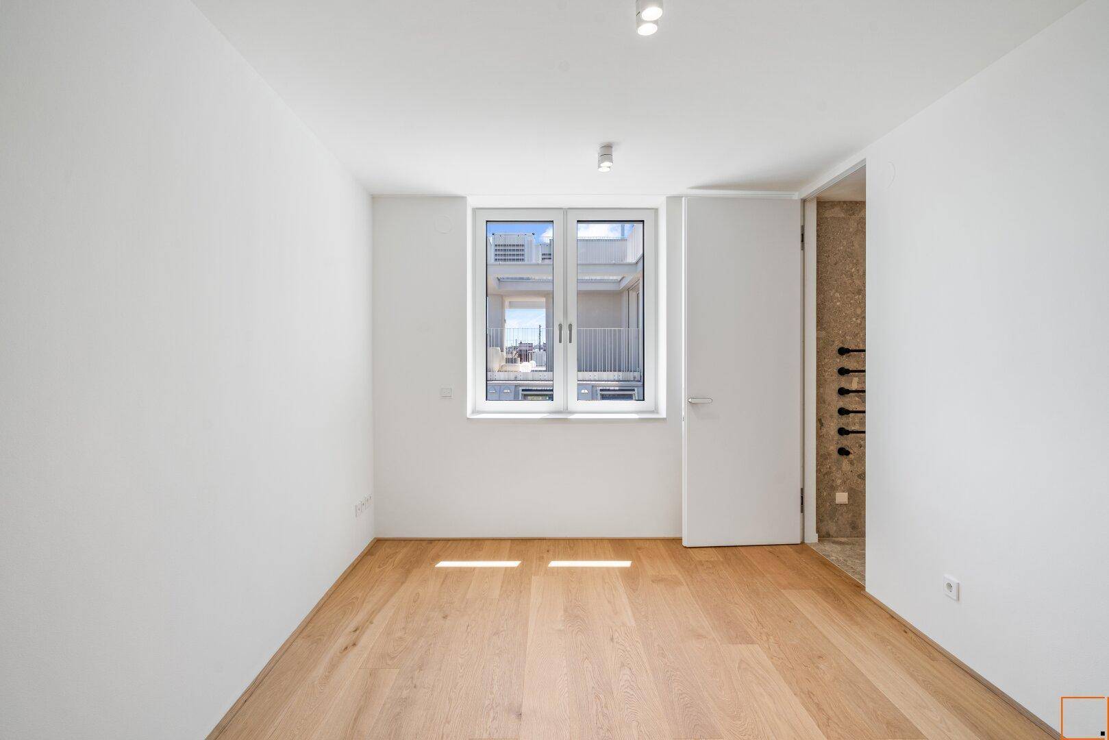 Leeres Zimmer mit Holzboden und Fenster, angrenzend an ein modernes Badezimmer.
