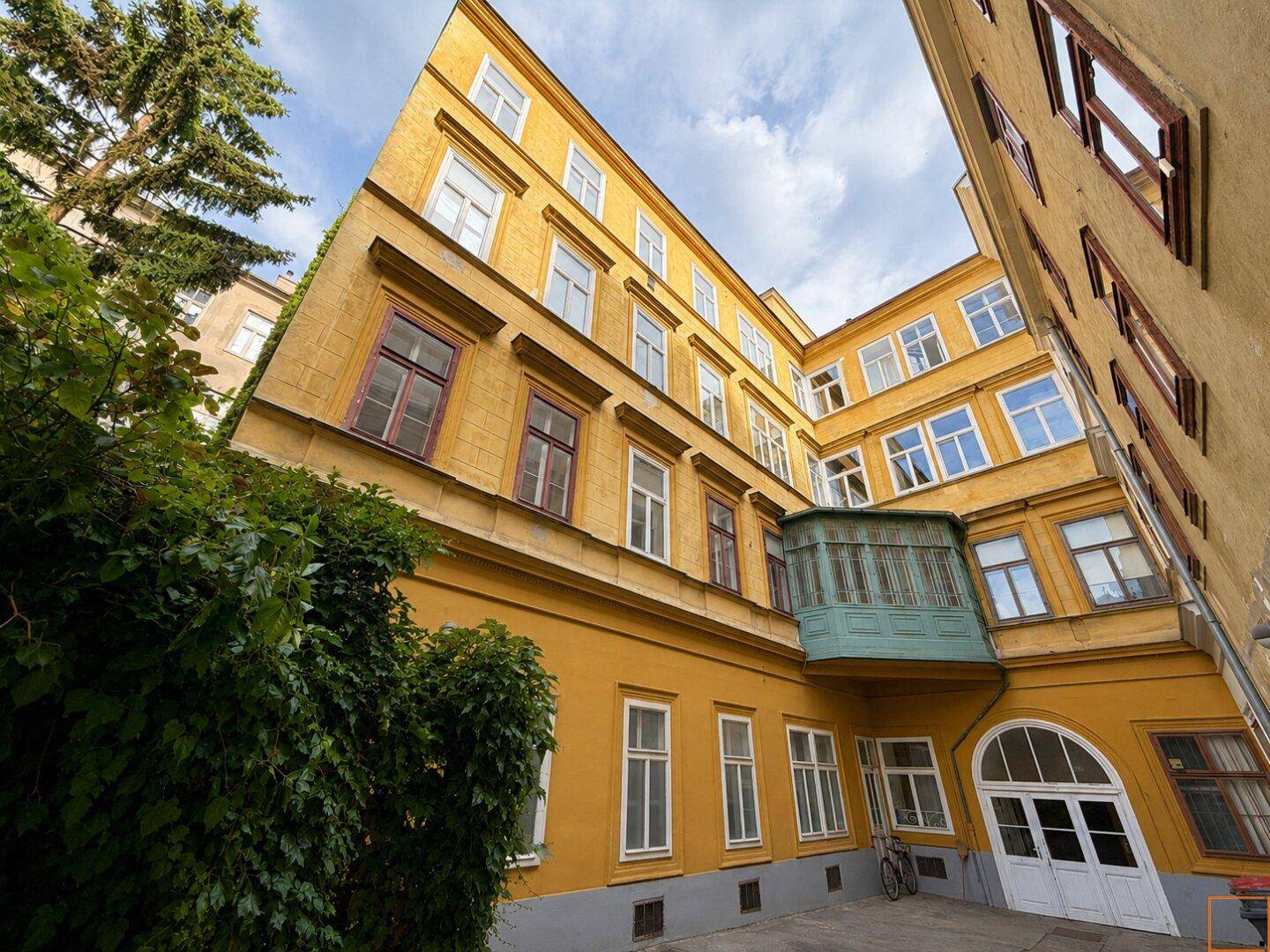 Außenansicht eines gelben Altbaus mit mehreren Stockwerken und einem kleinen, geschlossenen Balkon.