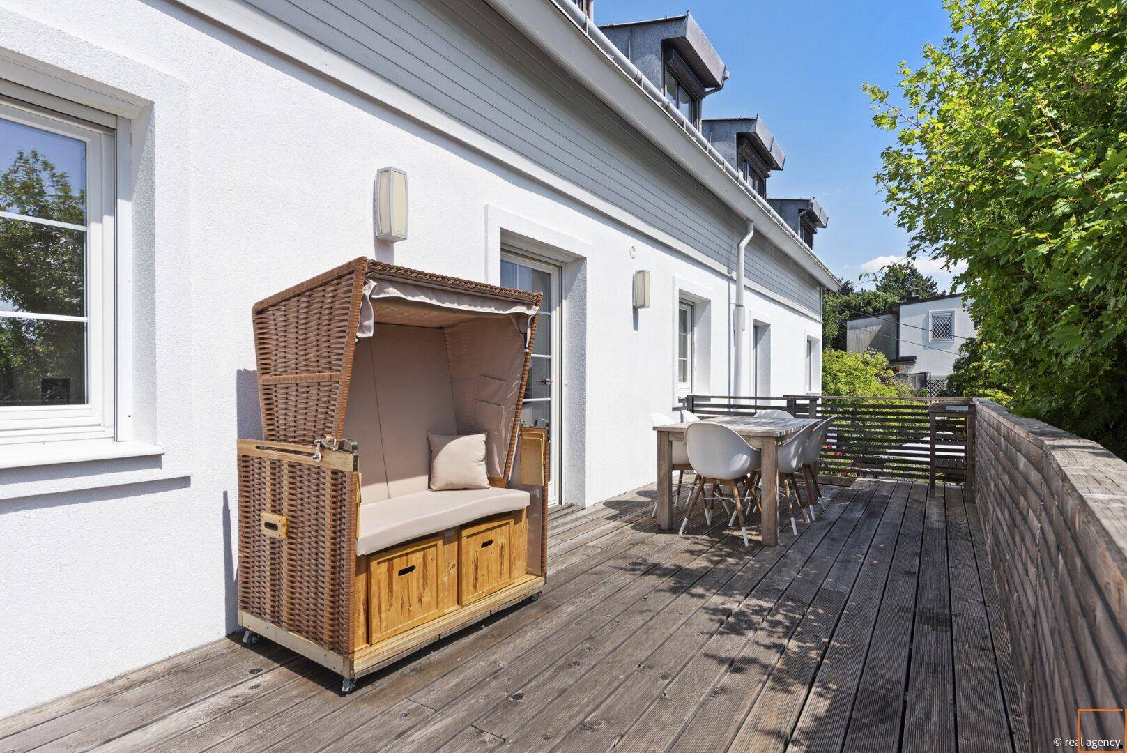 Geräumige Holzterrasse mit Strandkorb und Essbereich am weißen Haus.