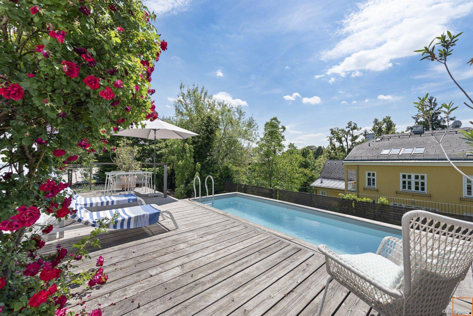 Sonnige Terrasse mit Pool, Sonnenliegen und blühenden Rosensträuchern mit Hausblick.