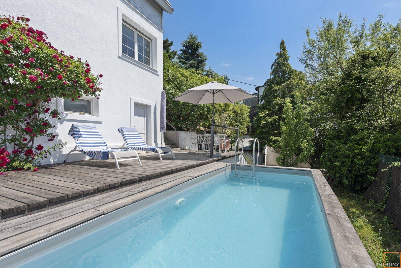 Hausfassade mit Holzterrasse, kleinem Pool und blühenden Rosensträuchern im Garten.
