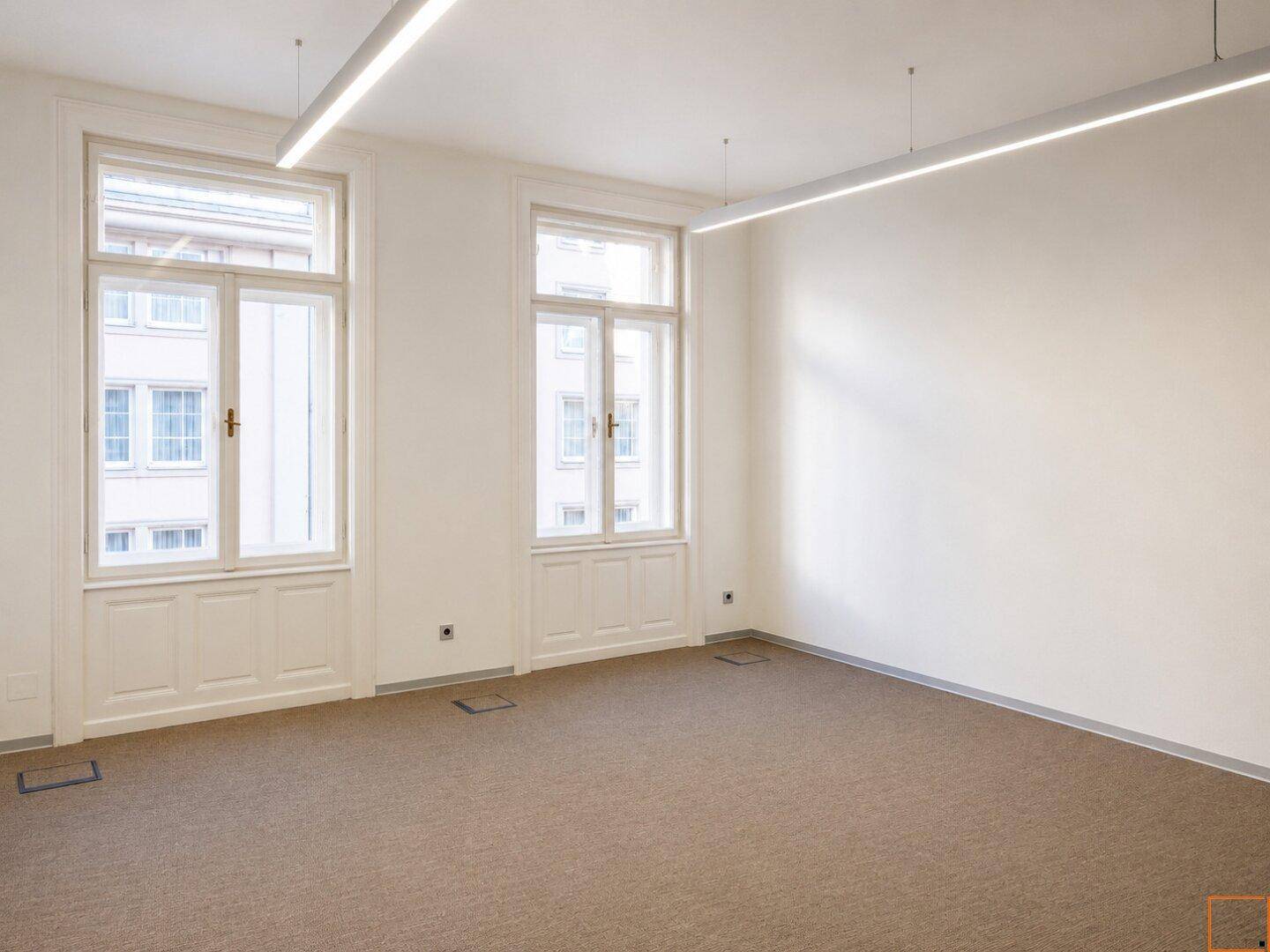 Heller Büroraum mit Teppichboden, hohen Fenstern und moderner, linearer Deckenbeleuchtung.