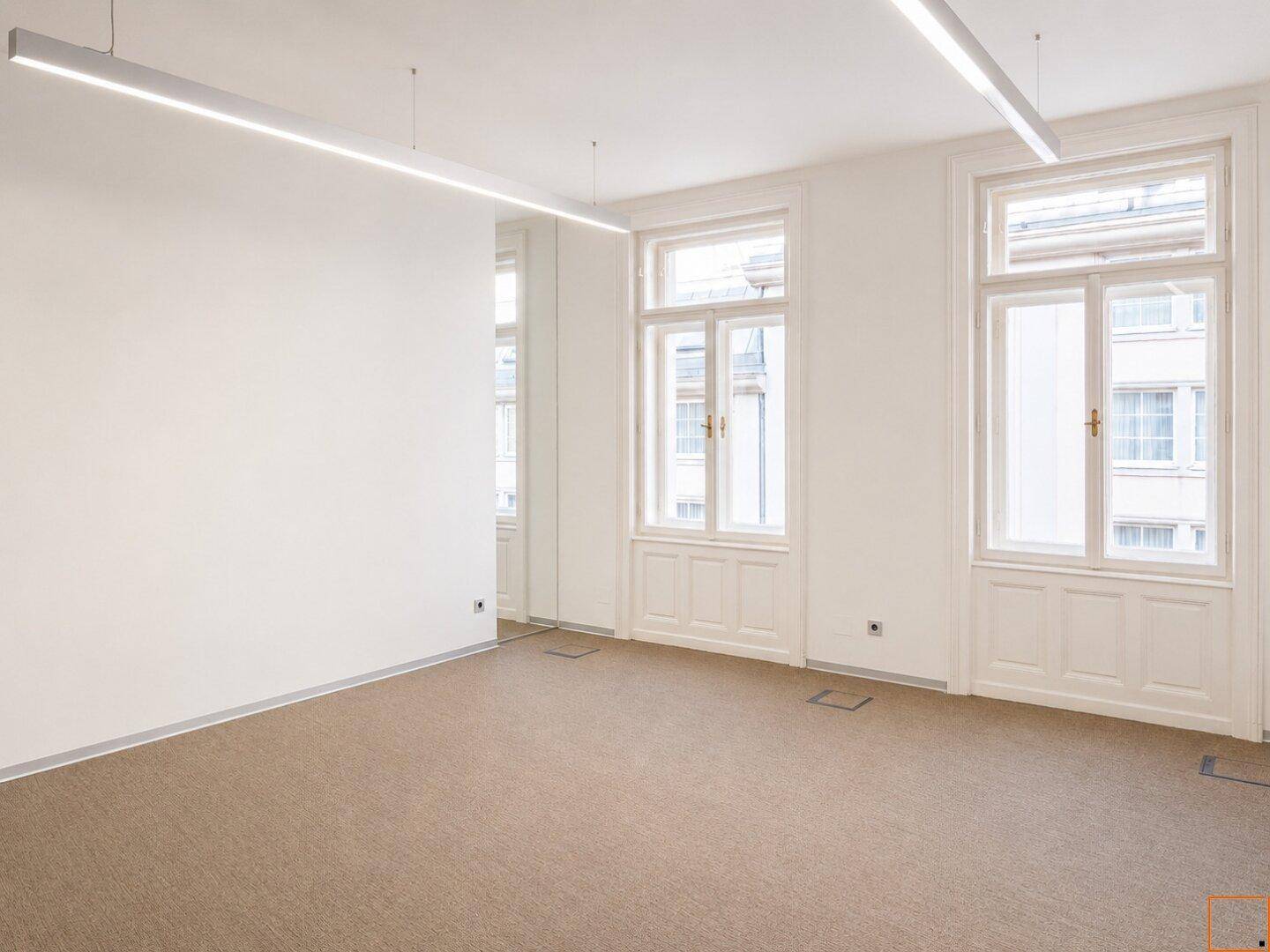 Leerer Büroraum mit Teppichboden, großen Fenstern und reflektierenden Wänden, die den Raum erweitern.