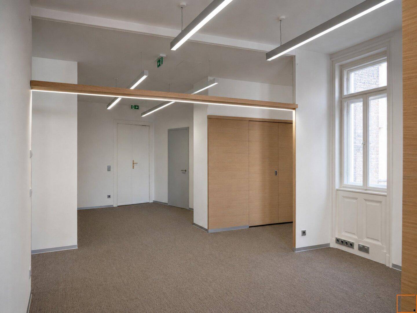 Geräumiger Büroraum mit Teppichboden, Holzelementen und einem großen Fenster für natürliches Licht.