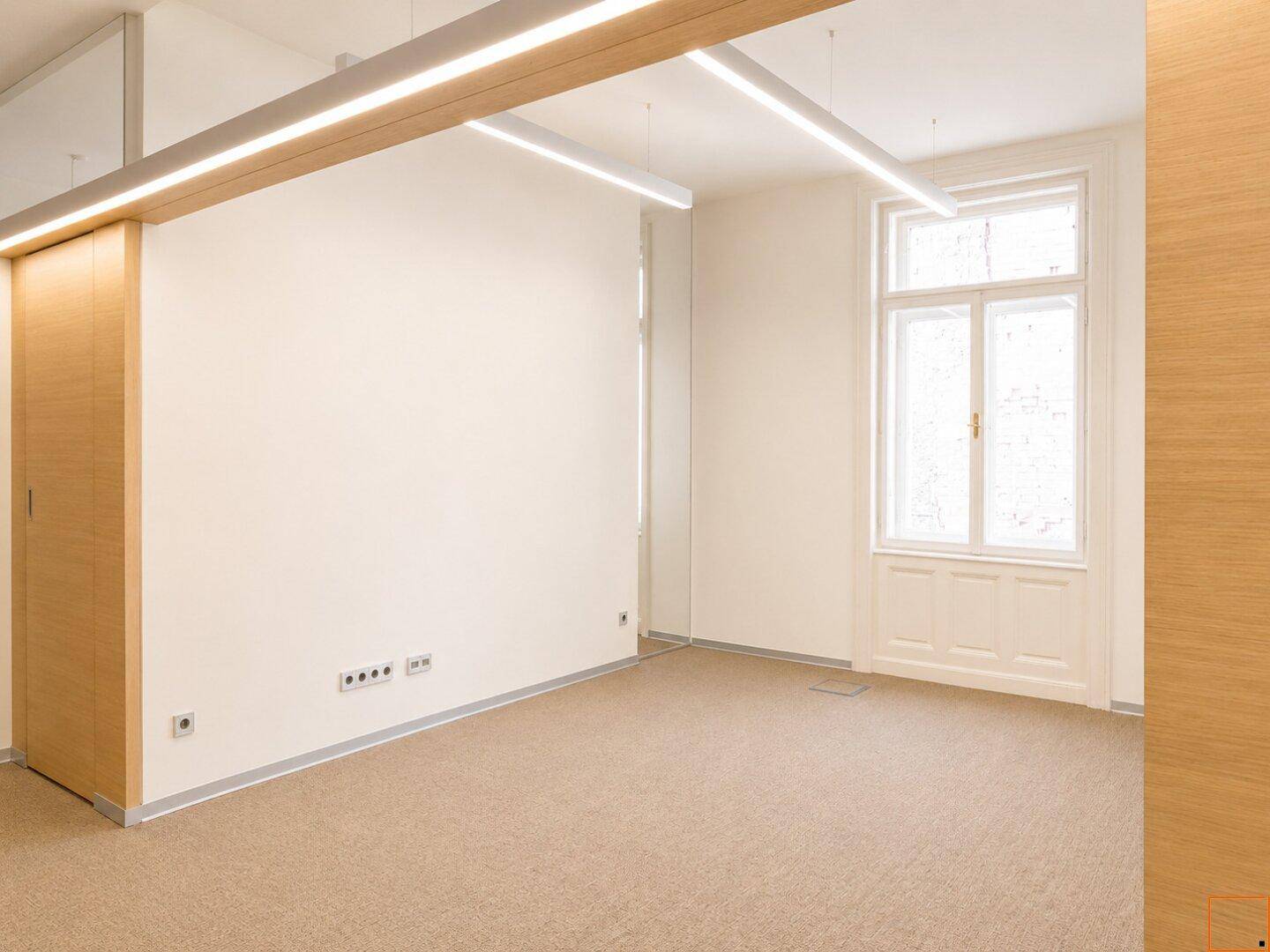 Büroraum mit Teppichboden, Holzelementen und einem großen Fenster, das viel Licht hereinlässt.