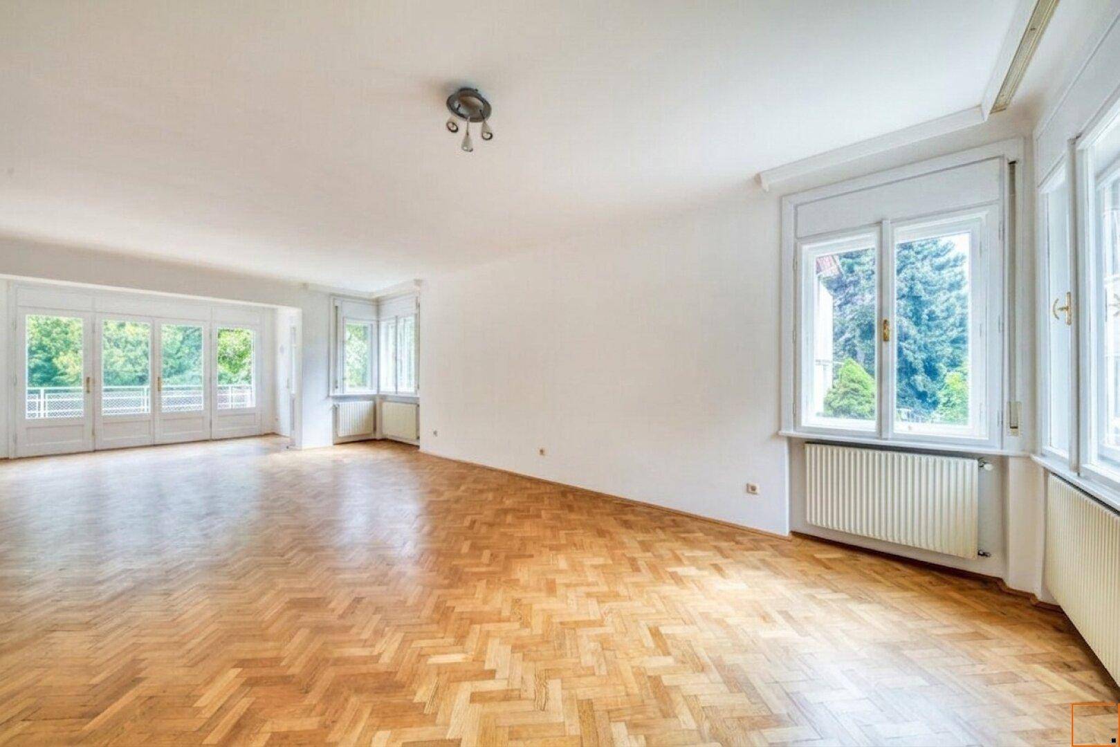 Weitläufiges Wohnzimmer mit Parkettboden und großen Fenstern, die viel Licht hereinlassen.