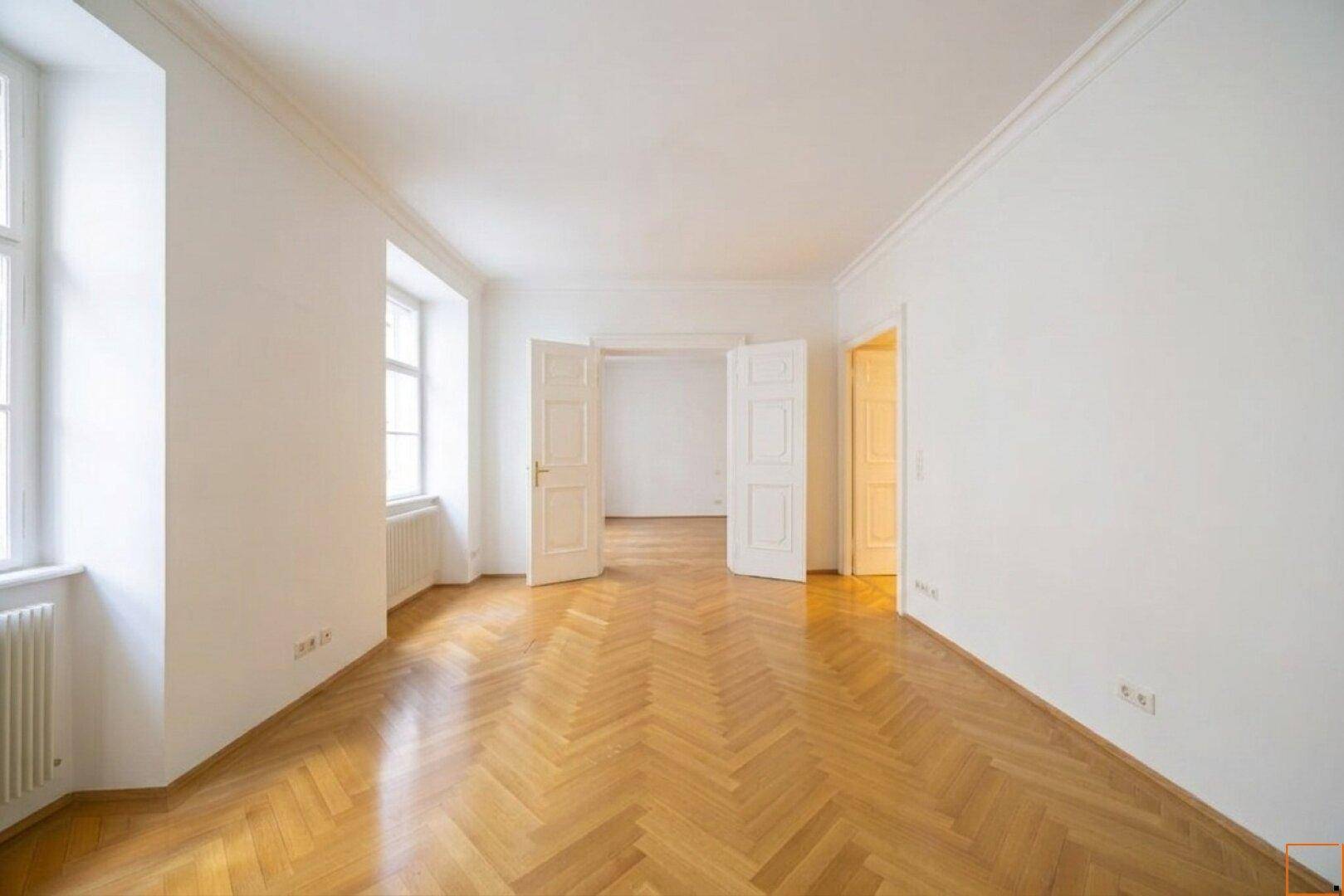 Geräumiger Wohnbereich mit Fischgrätparkett und eleganten Doppeltüren, die eine Verbindung zu weiteren Räumen schaffen.