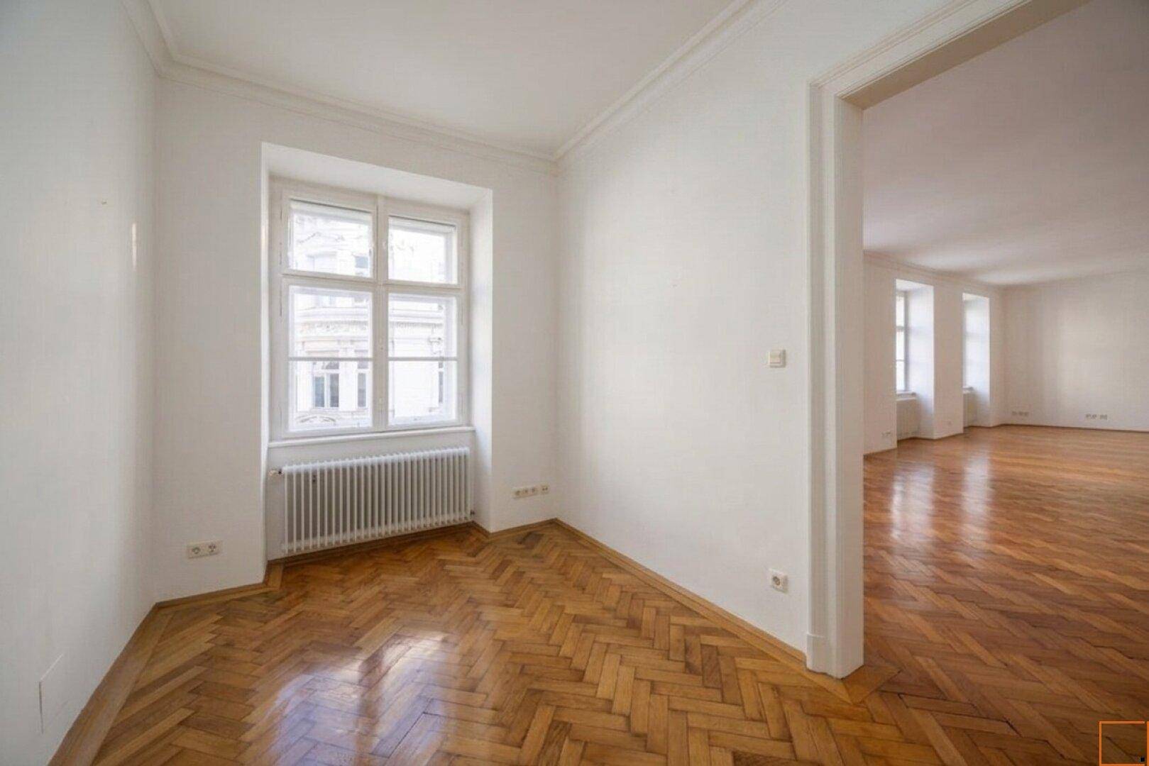 Kleinerer Raum mit Fischgrätparkett und einem Fenster, der durch eine offene Tür mit dem Hauptraum verbunden ist.