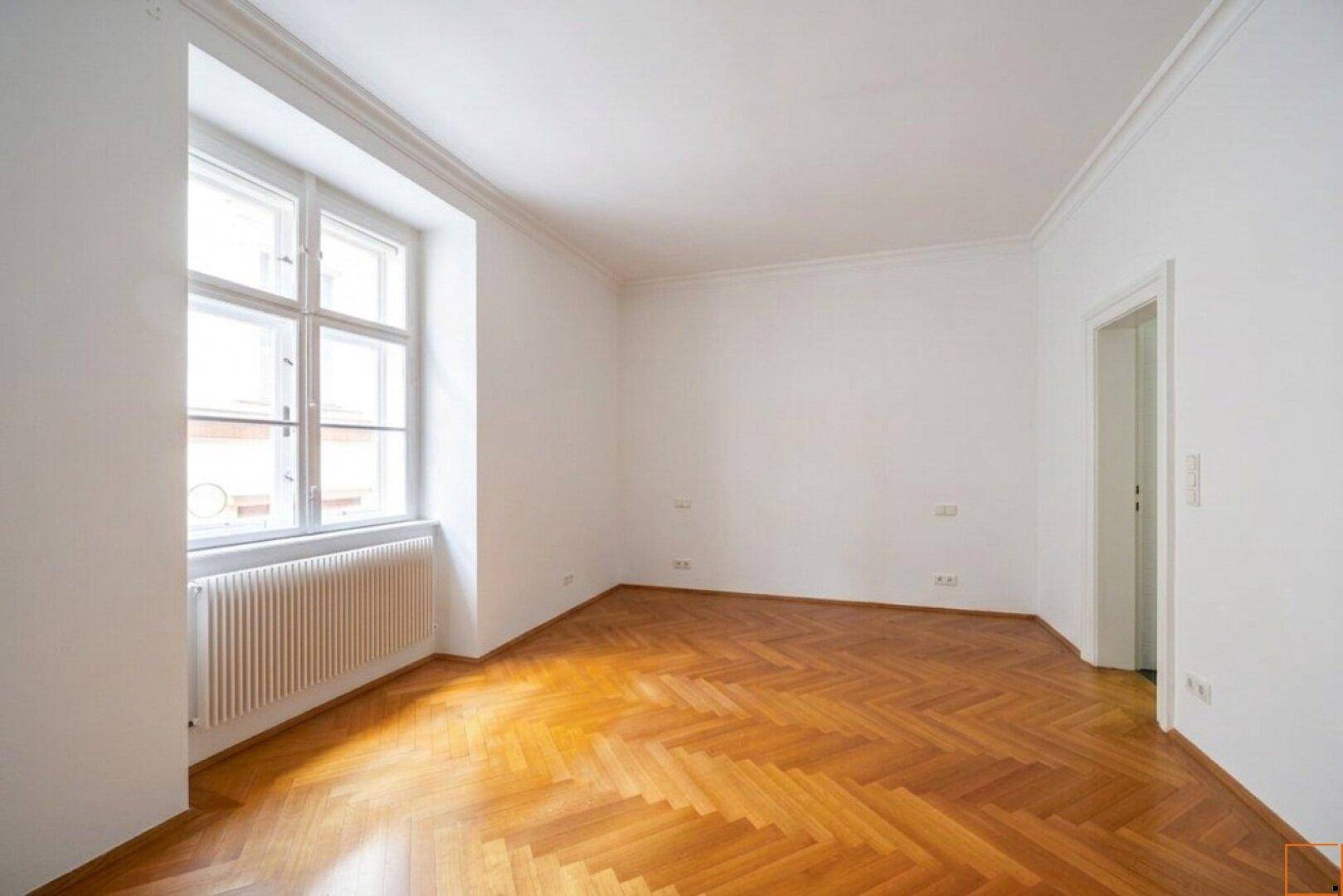 Heller Eckbereich eines Zimmers mit Fischgrätparkett und einem großen Fenster, das viel Licht hereinlässt.
