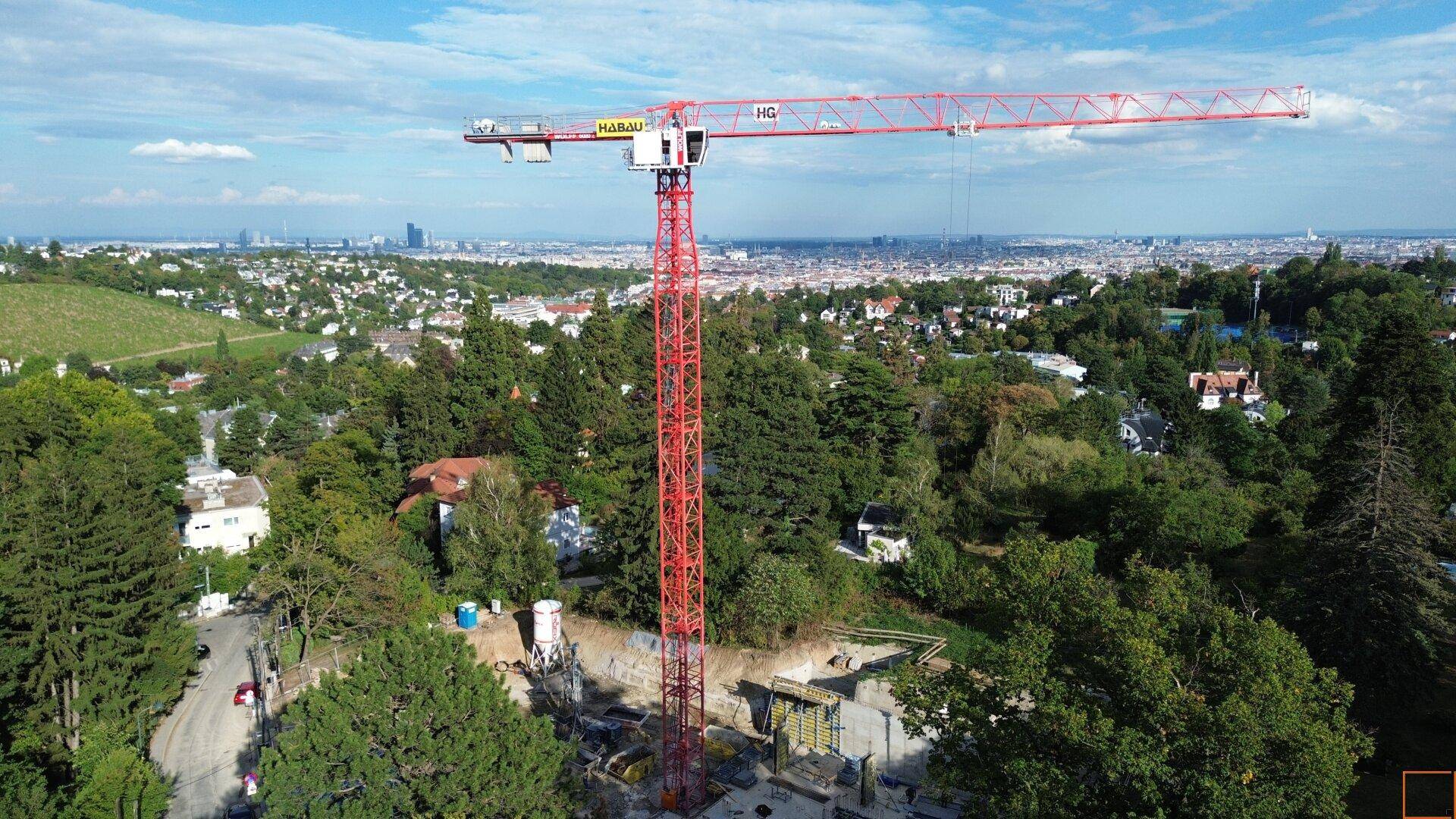 Baustelle mit Kran und Blick auf die Stadt, umgeben von grüner Hügellandschaft.