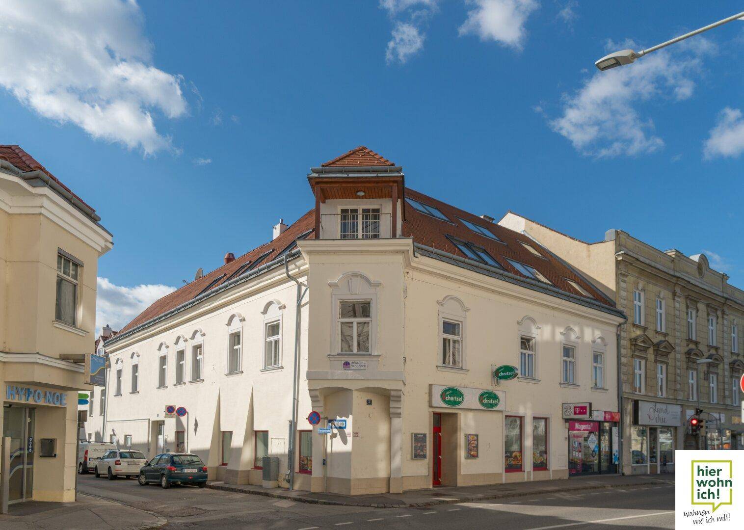 Historisches Eckgebäude mit Geschäften im Erdgeschoss und Wohnungen darüber, unter blauem Himmel.