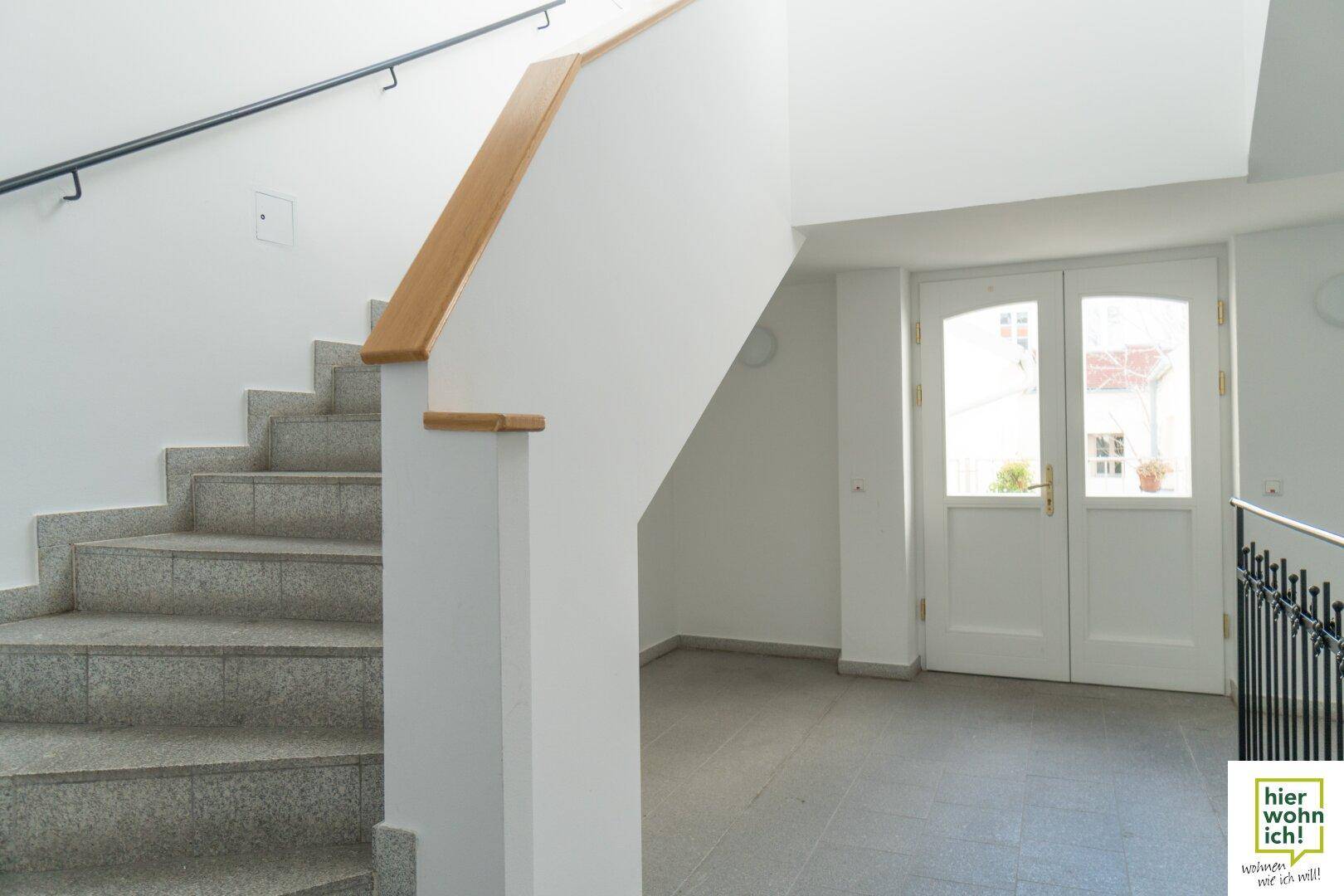 Heller Eingangsbereich mit Steintreppe, Holzgeländer und doppelter Glastür zum Hof.