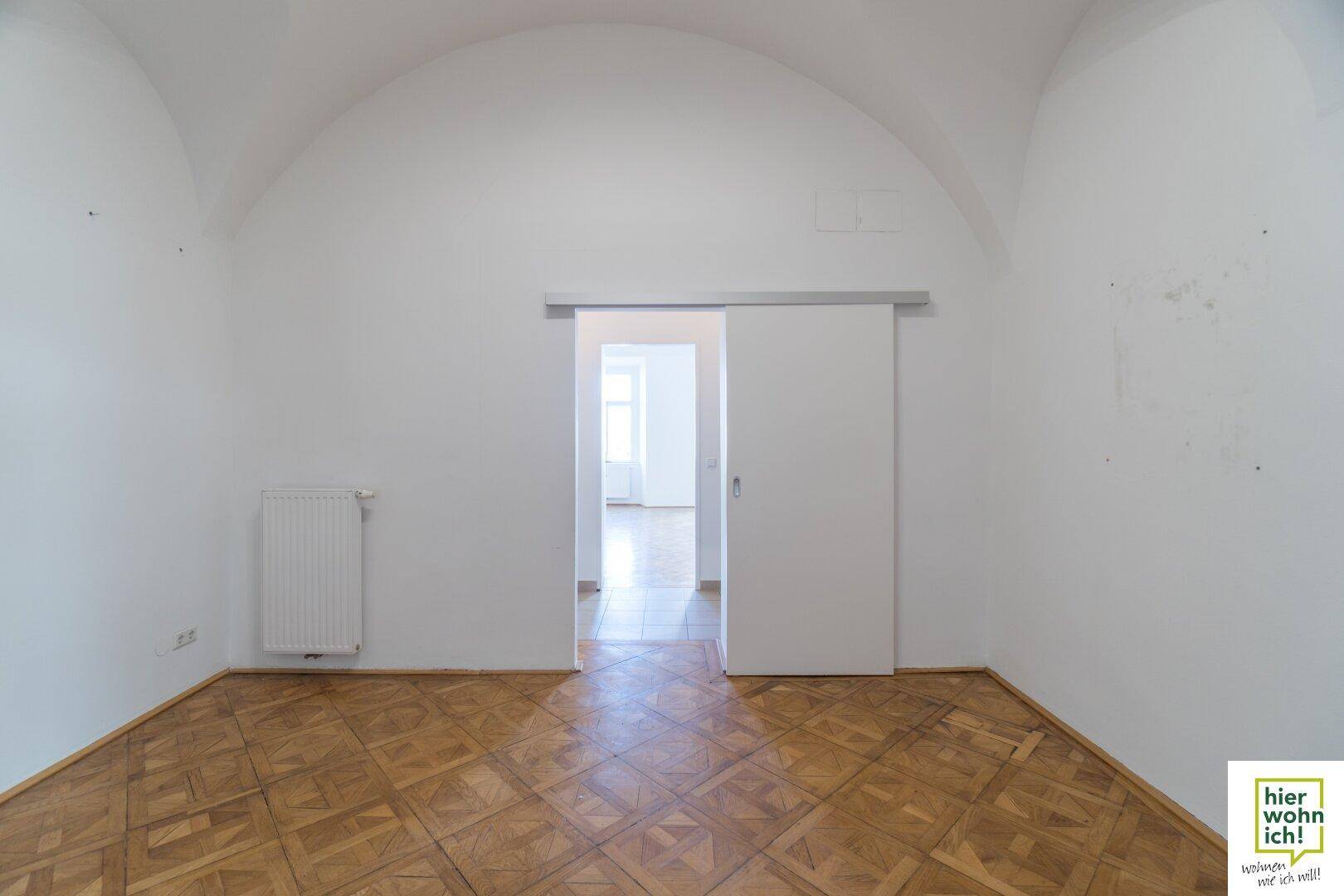 Raum mit Fischgrätparkett und einer Schiebetür, die zu einem weiteren Zimmer führt, unter einem Gewölbedecke.