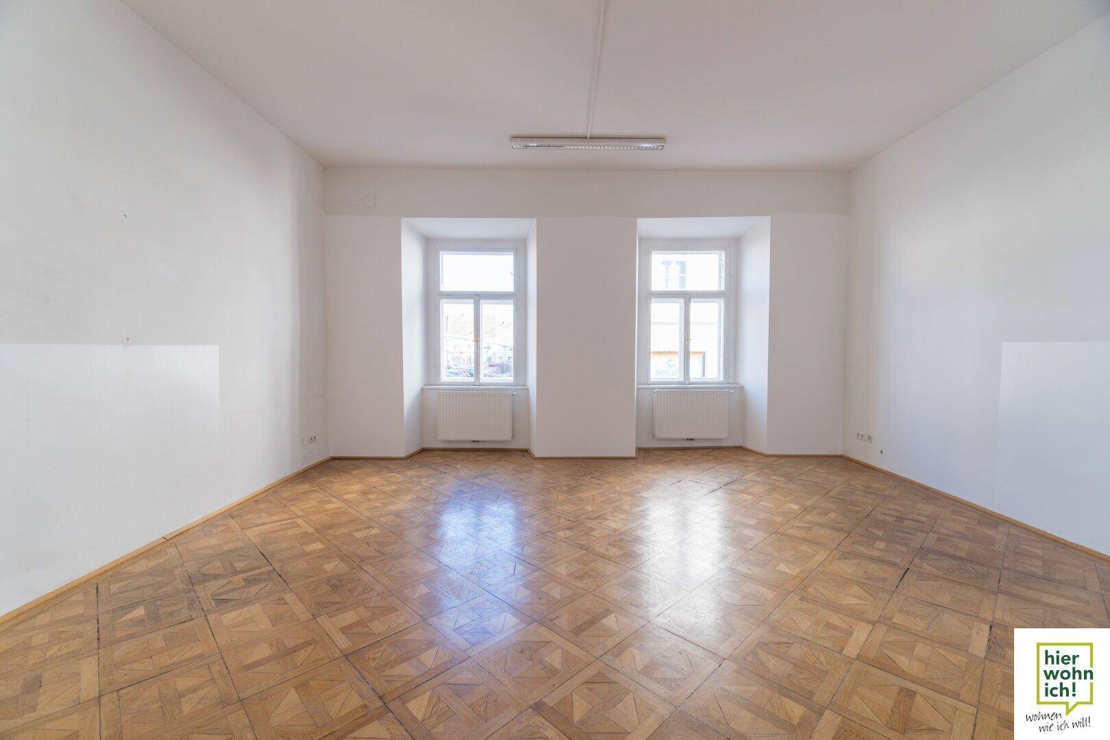 Geräumiger, heller Raum mit Fischgrätparkett und zwei großen Fenstern, ideal als Wohn- oder Arbeitsbereich.