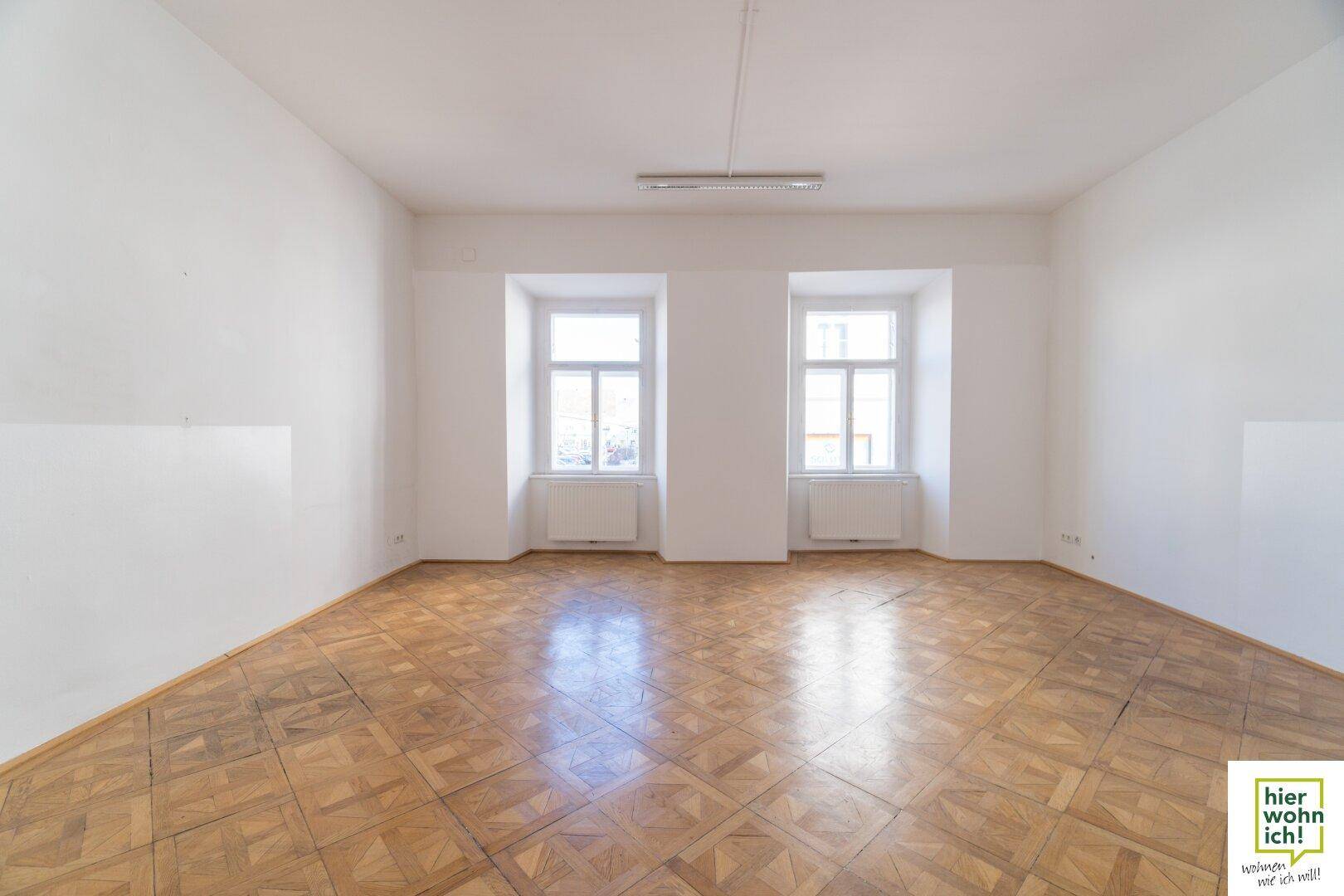 Geräumiger, heller Raum mit Fischgrätparkett und zwei großen Fenstern, ideal als Wohn- oder Arbeitsbereich.