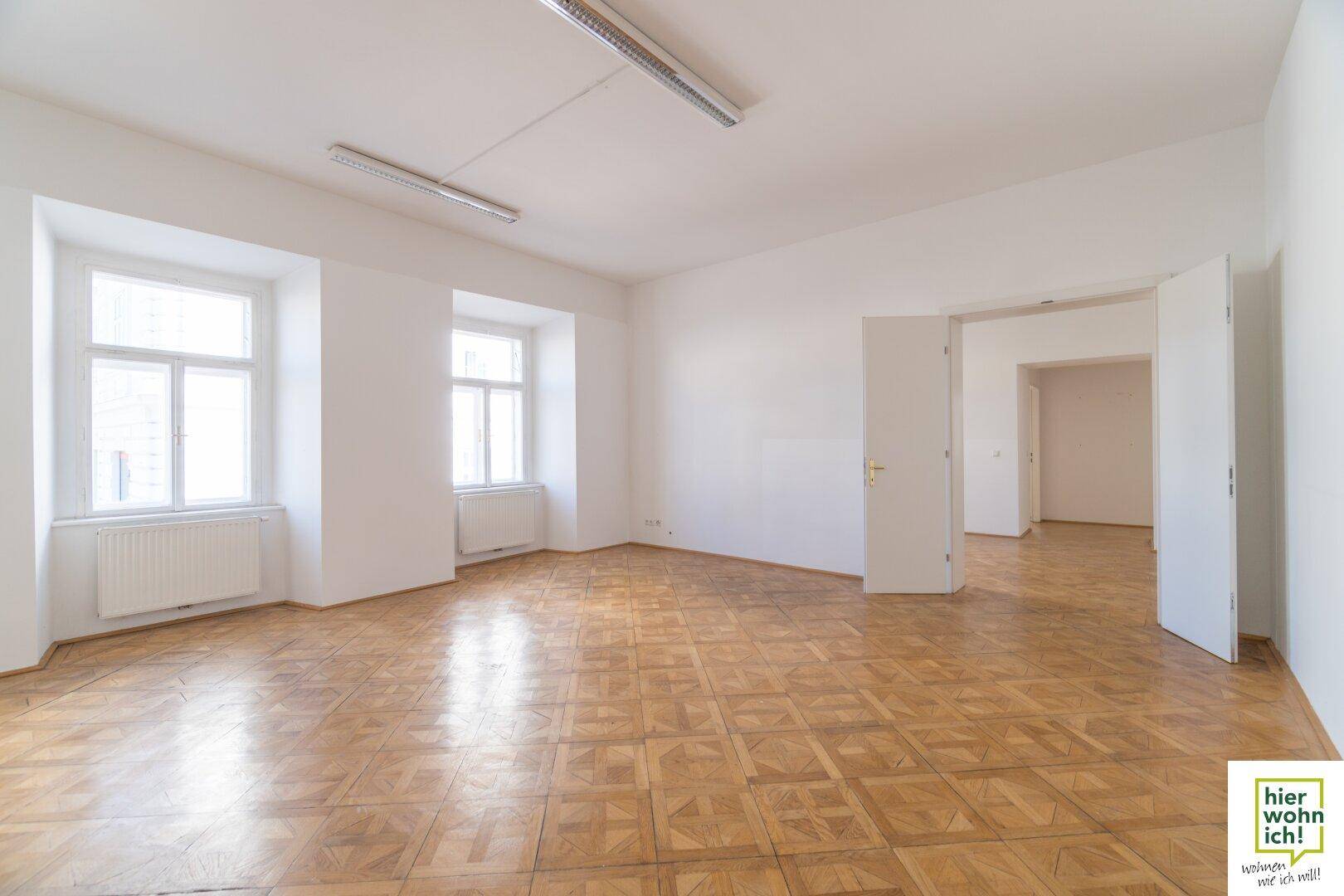 Geräumiger, heller Raum mit Fischgrätparkett und zwei großen Fenstern, ideal als Wohn- oder Arbeitsbereich.