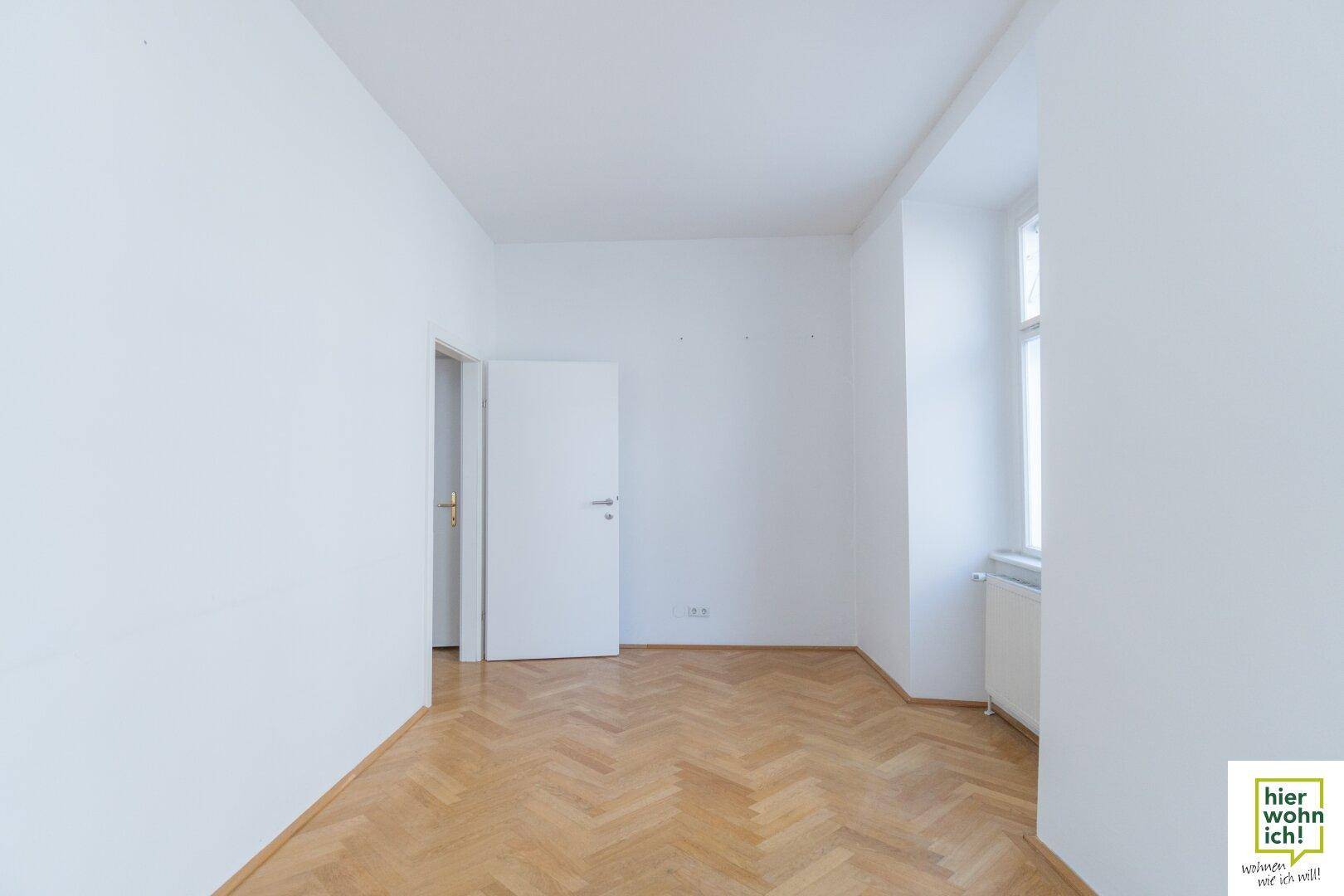 Heller Raum mit Fischgrätparkett und einer Tür, die zu einem weiteren Zimmer führt.