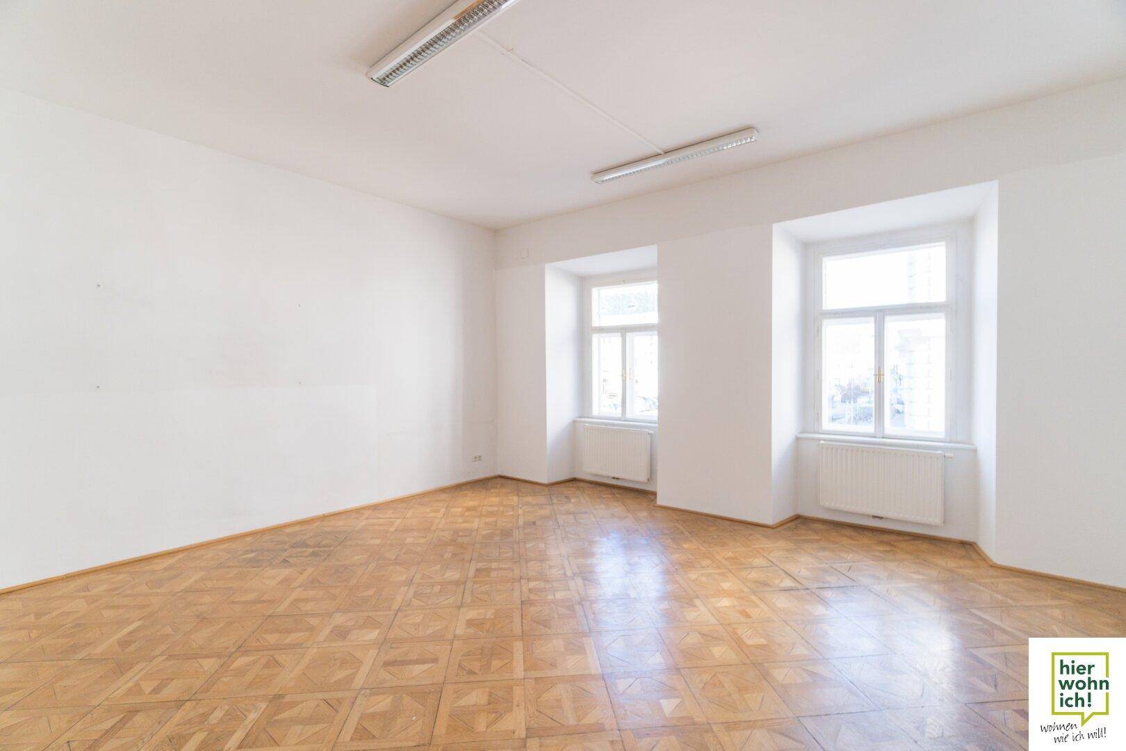 Geräumiger, heller Raum mit Fischgrätparkett und zwei großen Fenstern, ideal als Wohn- oder Arbeitsbereich.