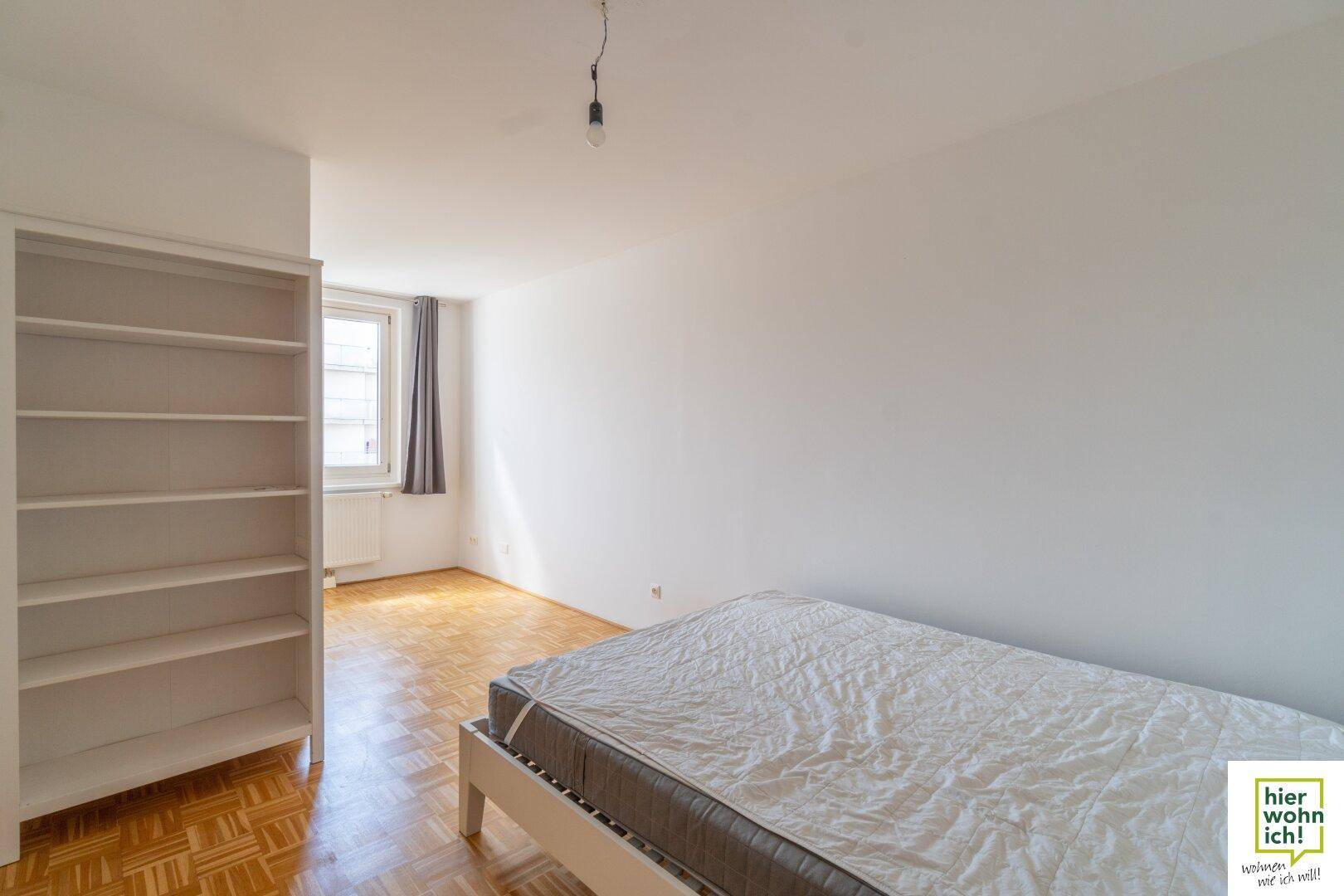 Schlafzimmer mit Parkettboden, Fenster und einem praktischen Bücherregal, hell und funktional.