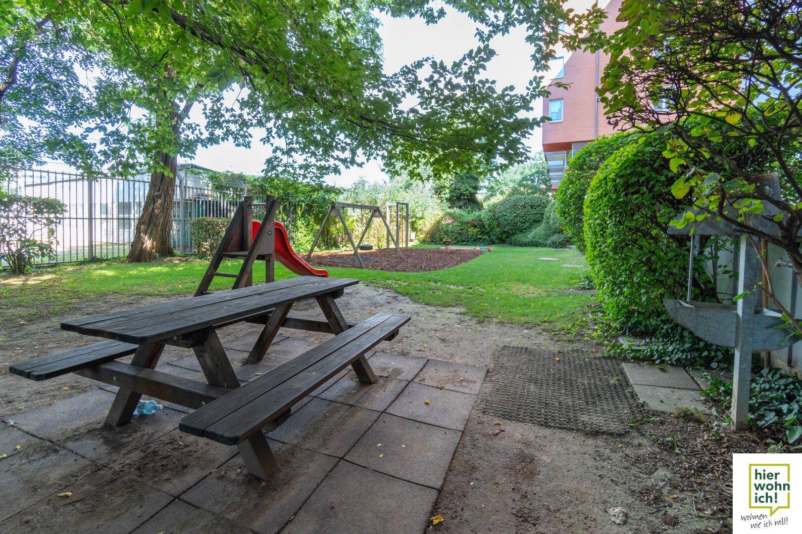 Gemeinschaftlicher Spielplatz mit Picknicktisch, Rutsche und Schaukeln unter schattenspendenden Bäumen.