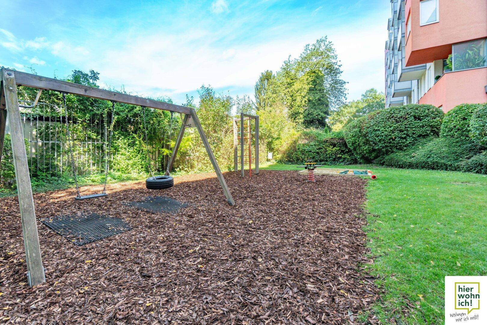 Großer Spielplatz mit Schaukeln und Sandkasten, umgeben von Grünflächen und Bäumen.
