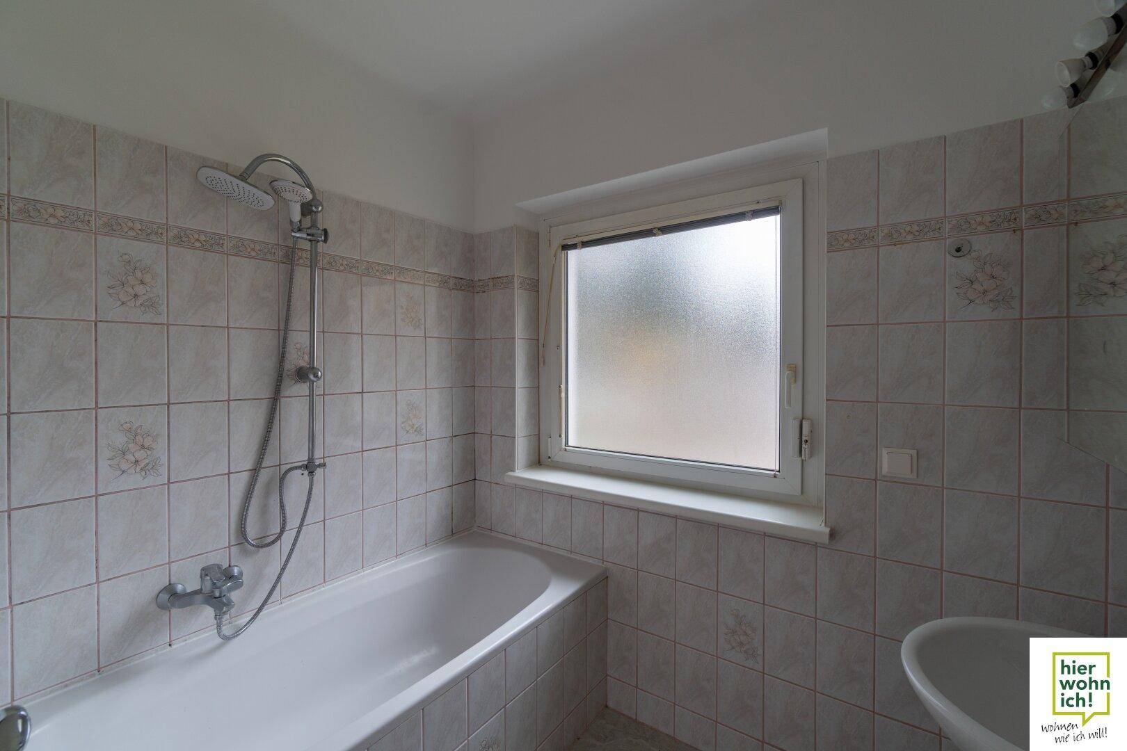 Badezimmer mit Badewanne, Duschfunktion und Fenster, komplett gefliest in hellen Tönen.