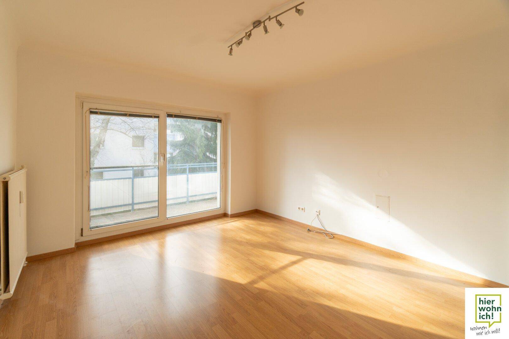 Helles Wohnzimmer mit Parkettboden und Zugang zu einem Balkon, durchflutet von natürlichem Sonnenlicht.
